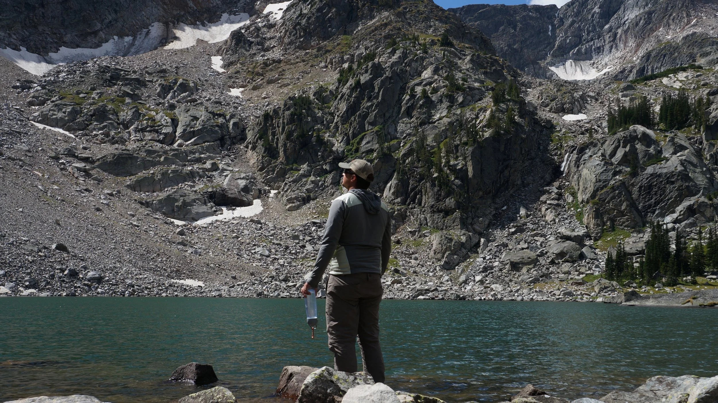 Person standing by a mountain lake, holding a water filter, surrounded by rocky terrain and snow patches on steep cliffs.
