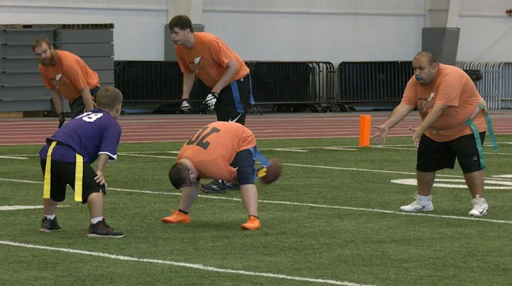 Special needs football game with players and coaches on an indoor field, one player is in a wheelchair.