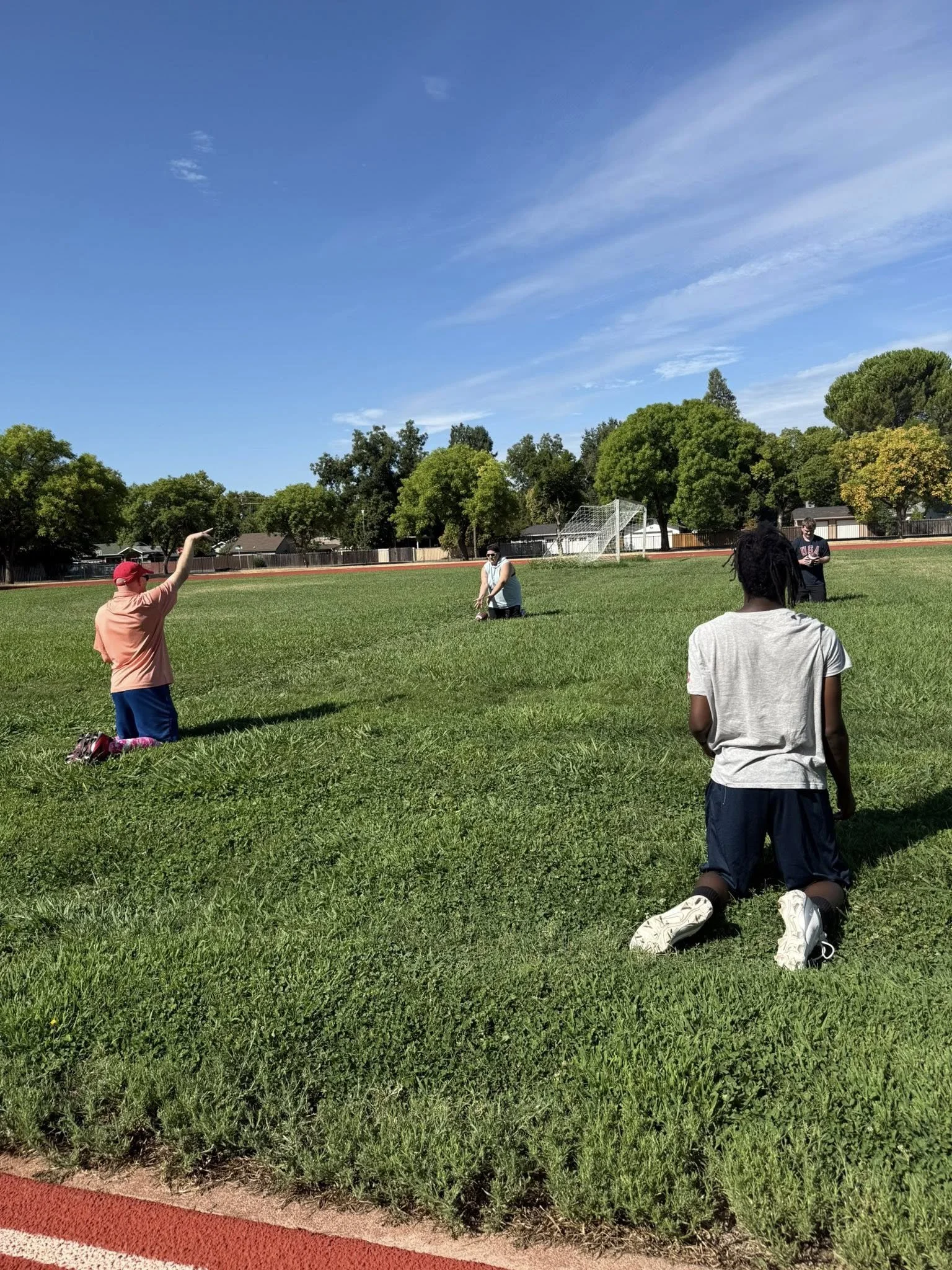 Four people are on a grassy field on a sunny day, with trees and sports facilities in the background. One person is kneeling, another is standing with their arm raised, and two more are standing in the distance.
