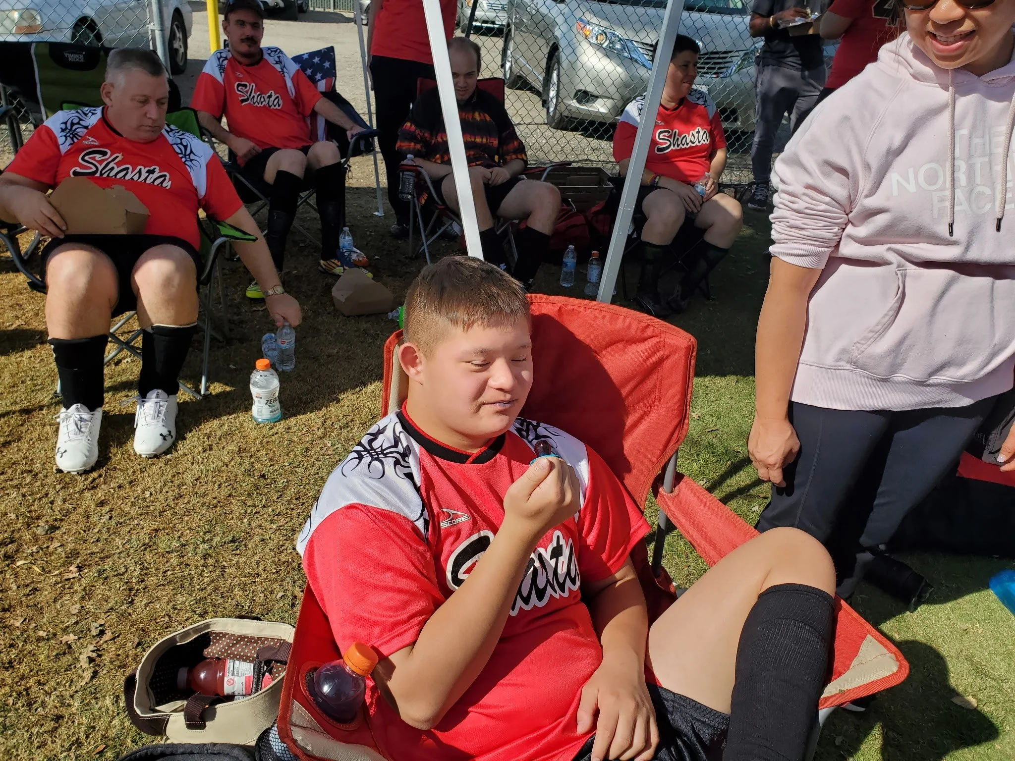 A young boy in a red sports uniform sitting in a chair, holding an ice cube, with his eyes closed. Behind him, several people are seated, some wearing matching red sports jerseys, at an outdoor event on grassy ground, with cars and a chain-link fence in the background.