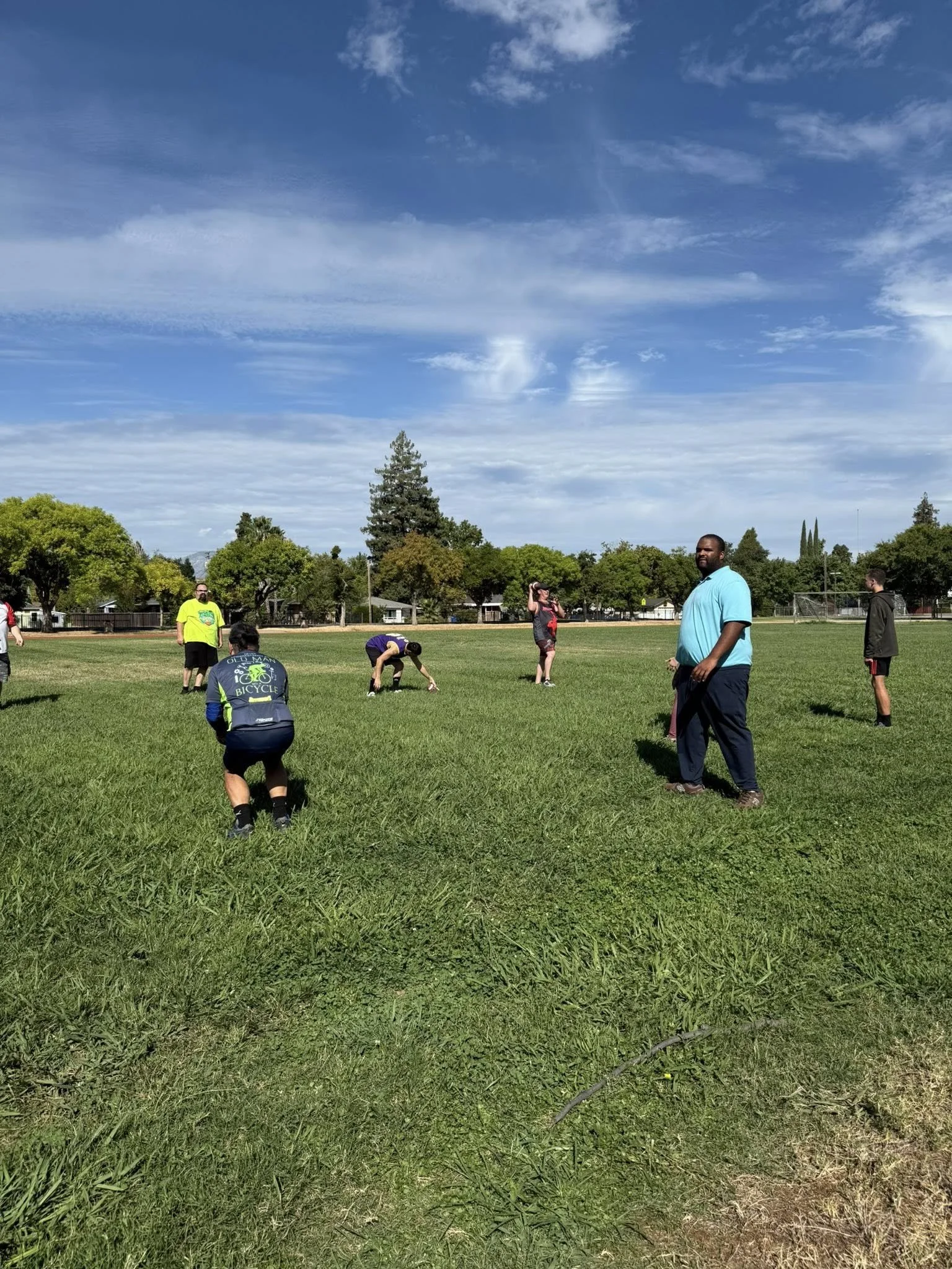 People standing and sitting on a grassy field, some stretching and preparing, with trees and houses in the background under a partly cloudy blue sky.