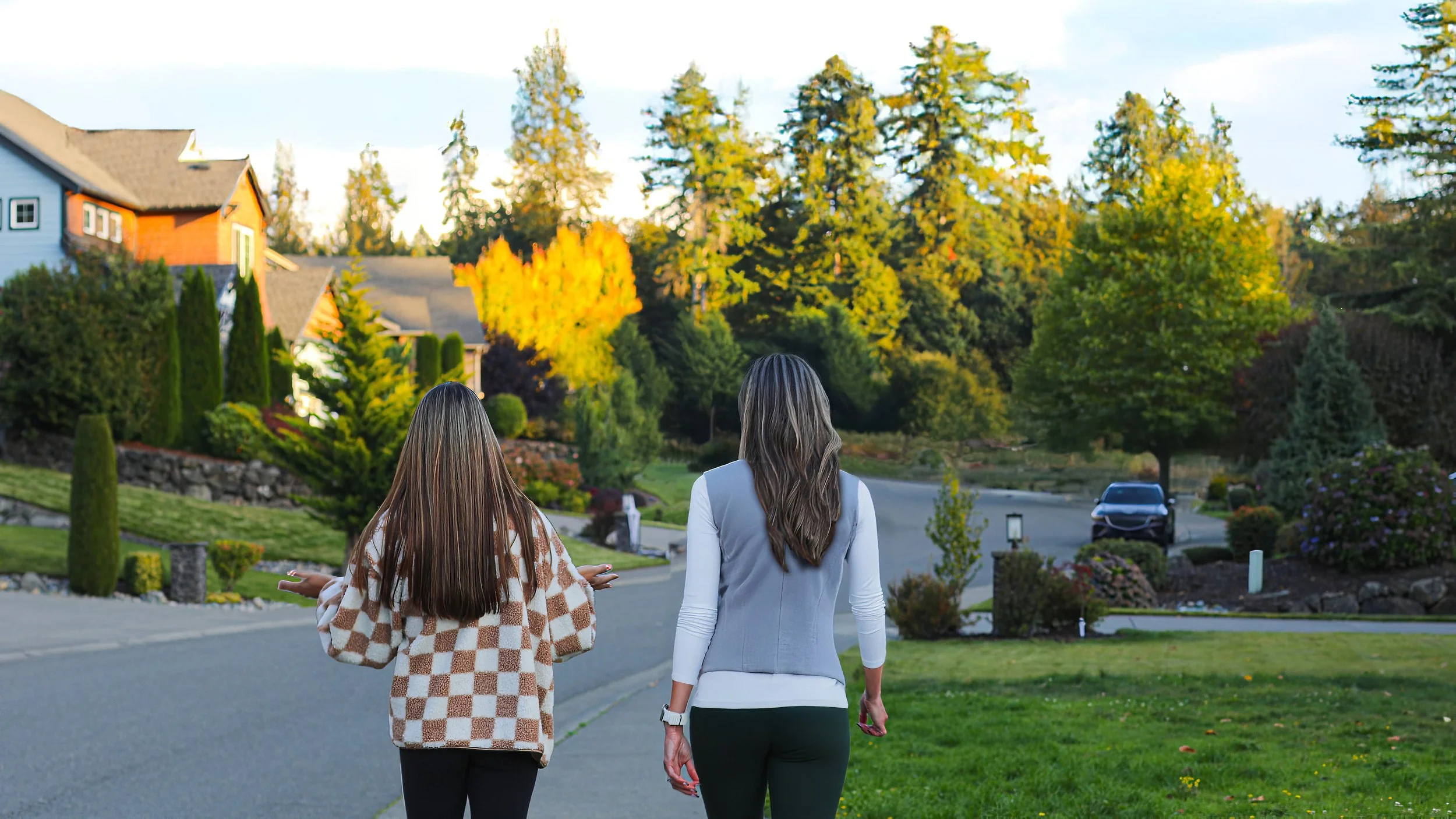 Two women walking down a suburban street with landscaped yards and trees in autumn colors.