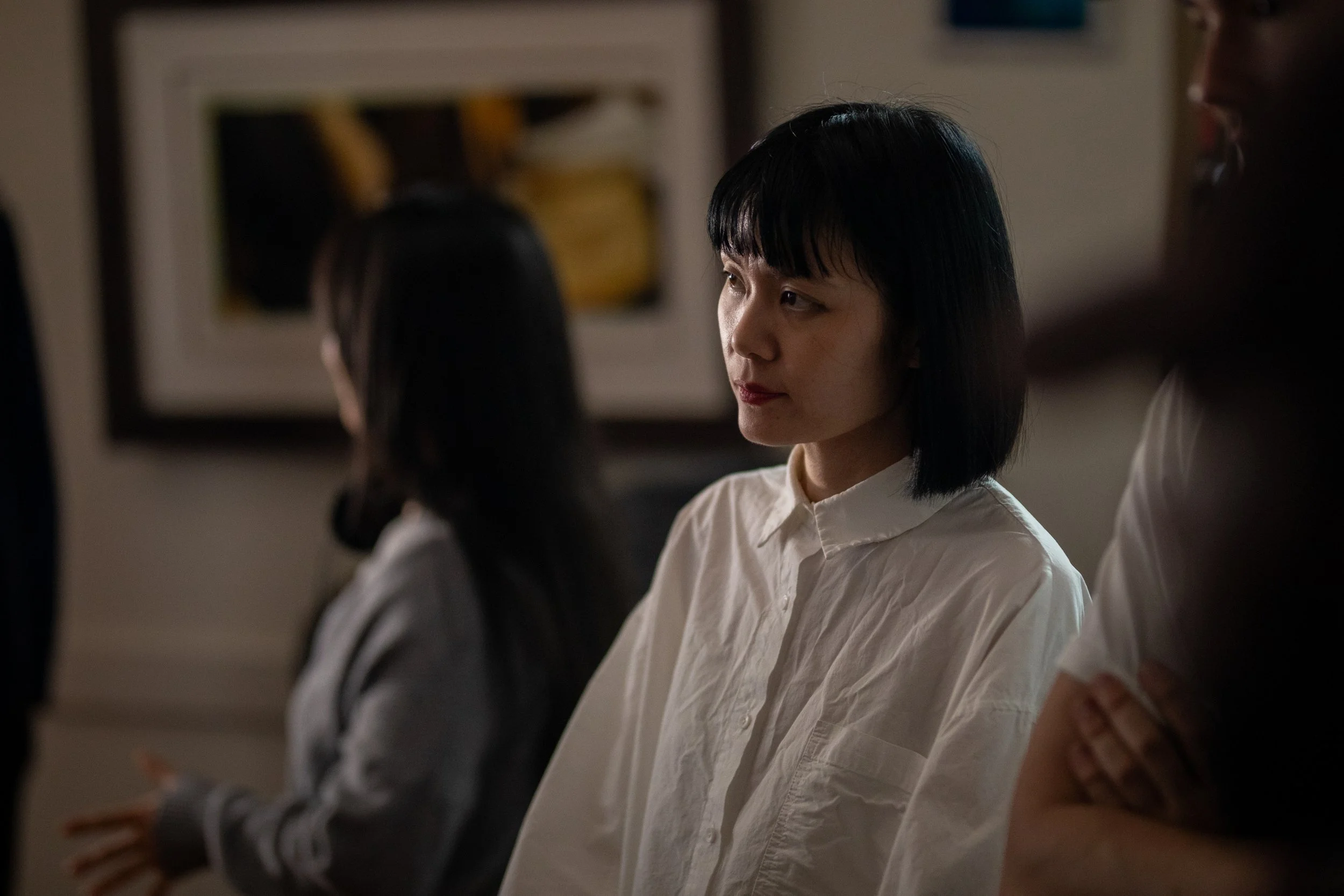 People in a dimly lit room, one woman in focus wearing a white shirt, others blurred in the background.