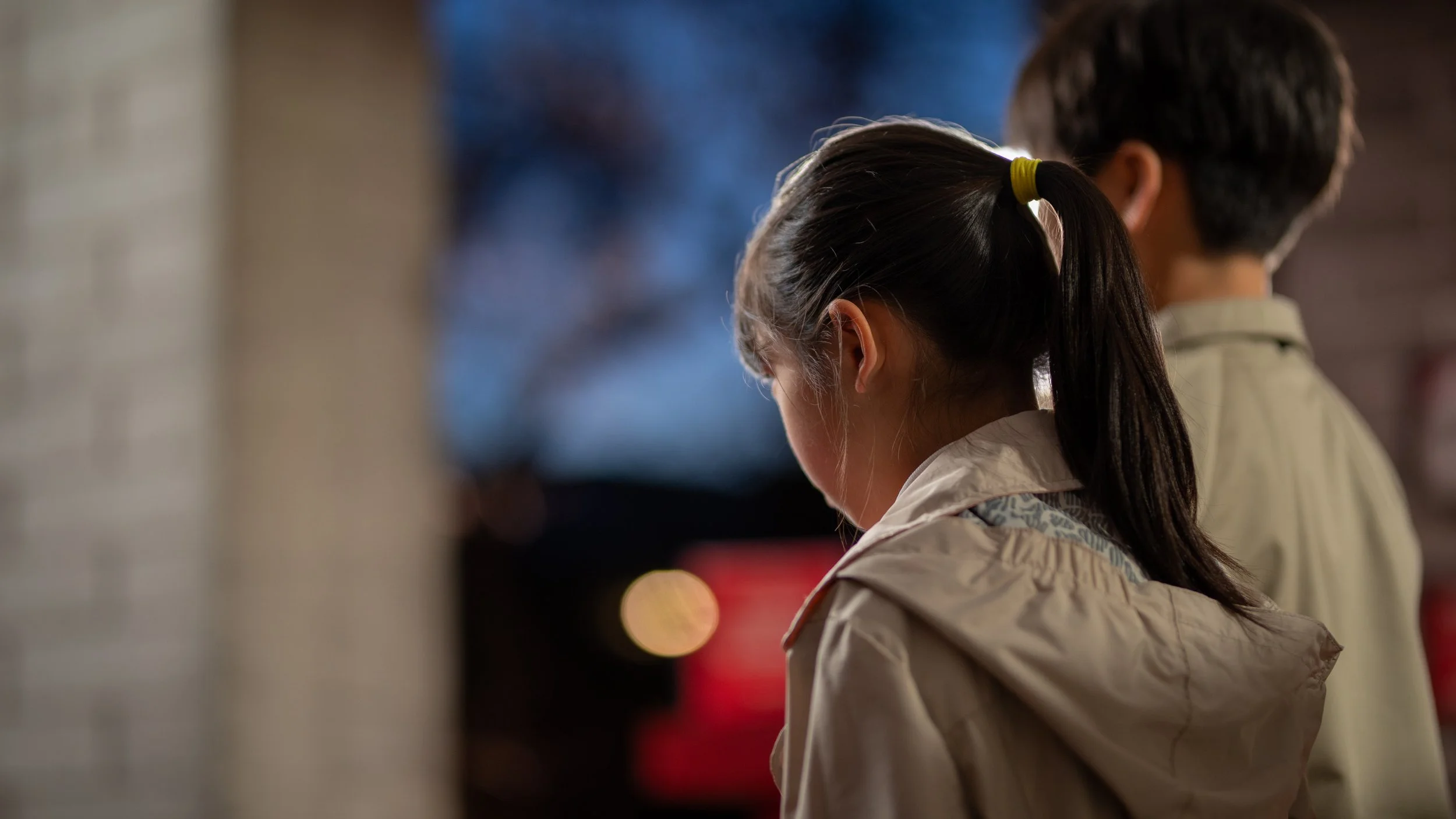 Rear view of two children standing together, one with a ponytail and the other with short hair, wearing light-colored jackets, with a blurred background.