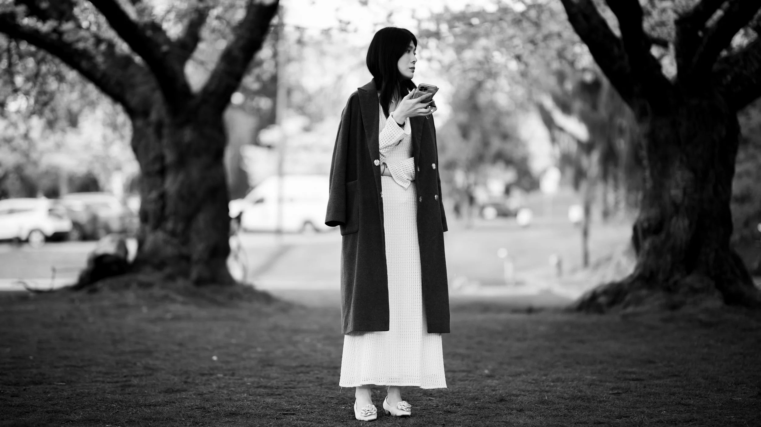 Black and white image of a woman standing in a park, wearing a long coat and holding a smartphone. Large trees and parked cars are visible in the background.