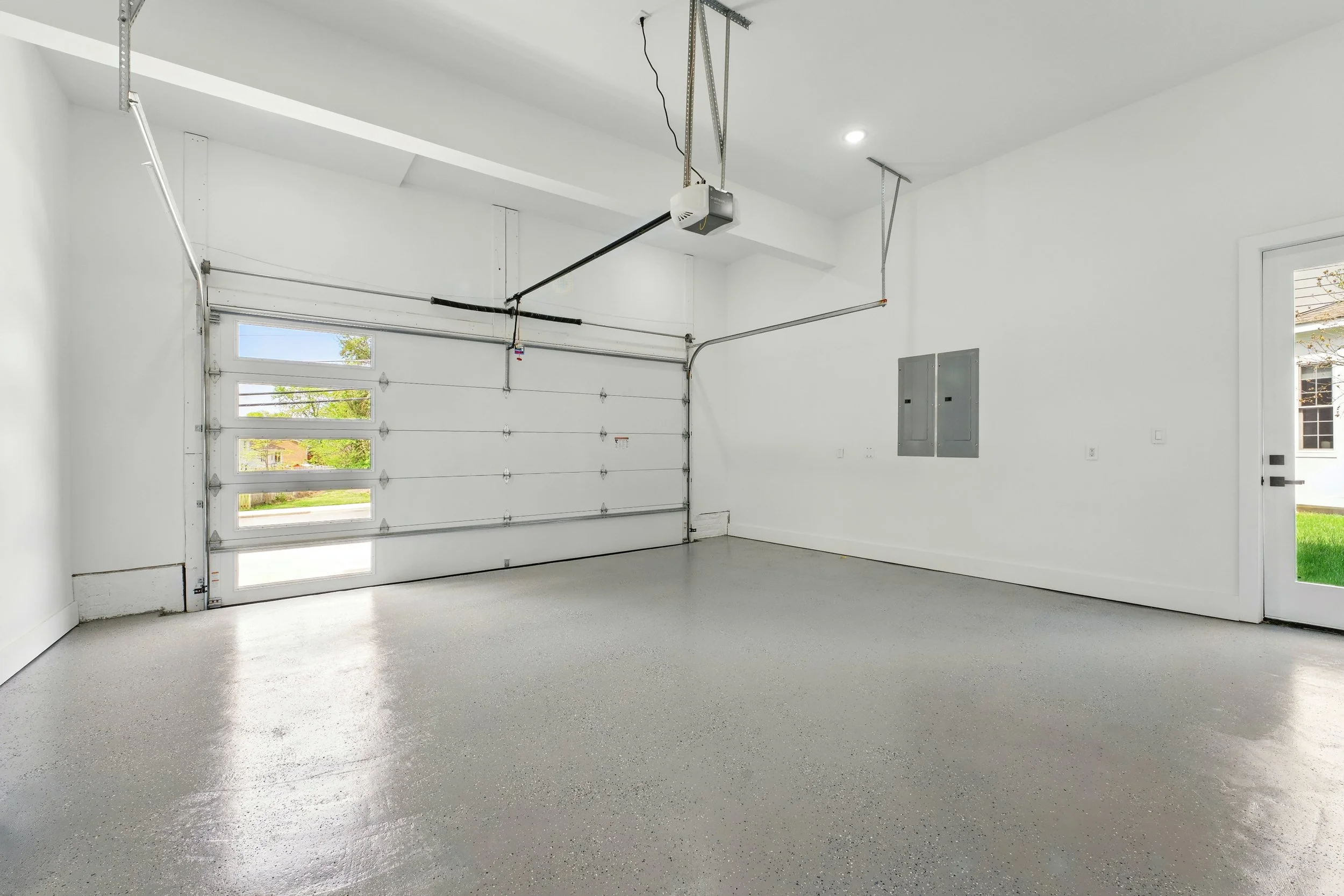 Empty clean garage with a white garage door, concrete flooring, and a door leading outside to grass. The garage has electrical panels on the wall and an automatic garage door opener.