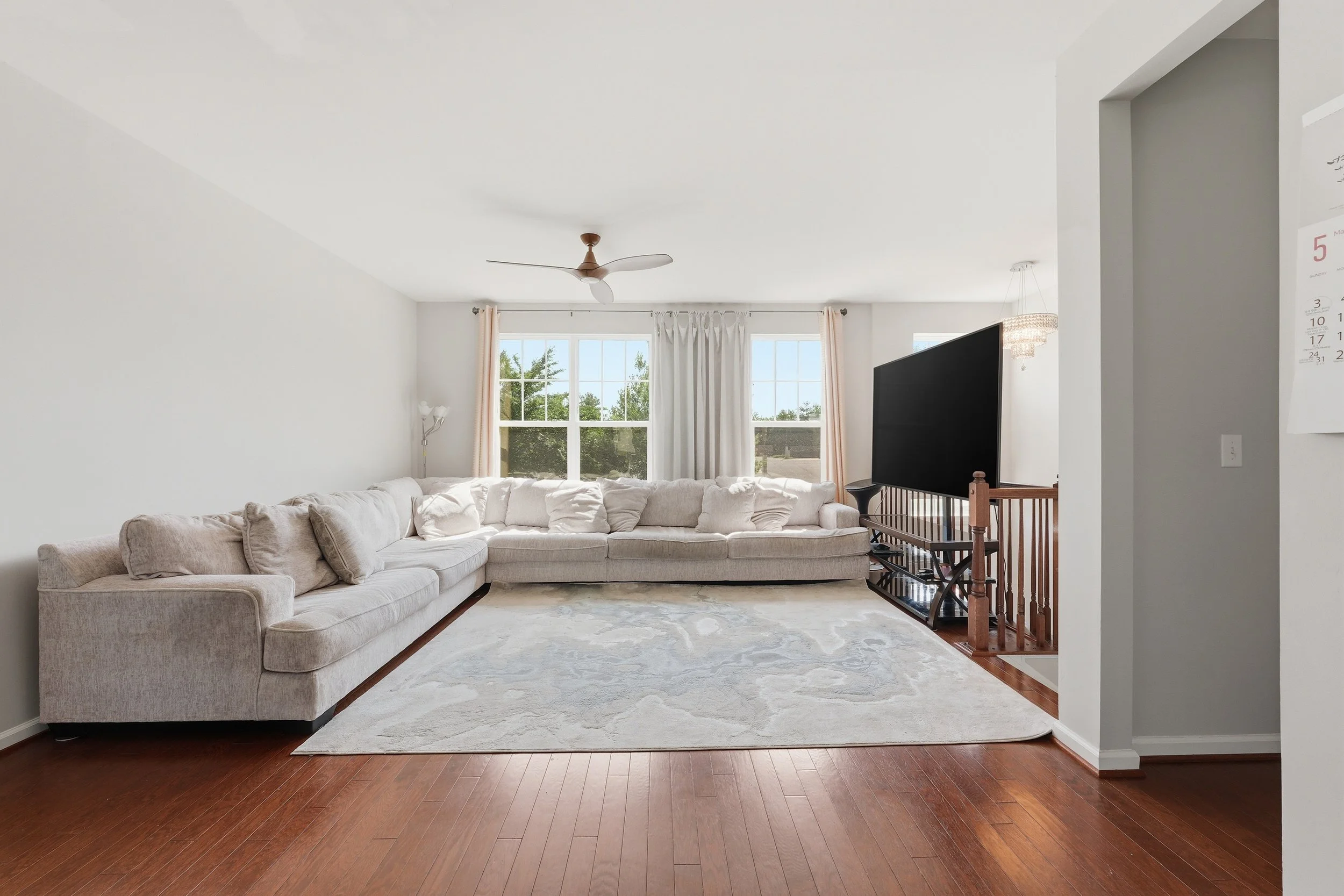 Living room with a large pale sectional beneath two windows, a marbled gray area rug, a ceiling fan and dark hardwood floors.