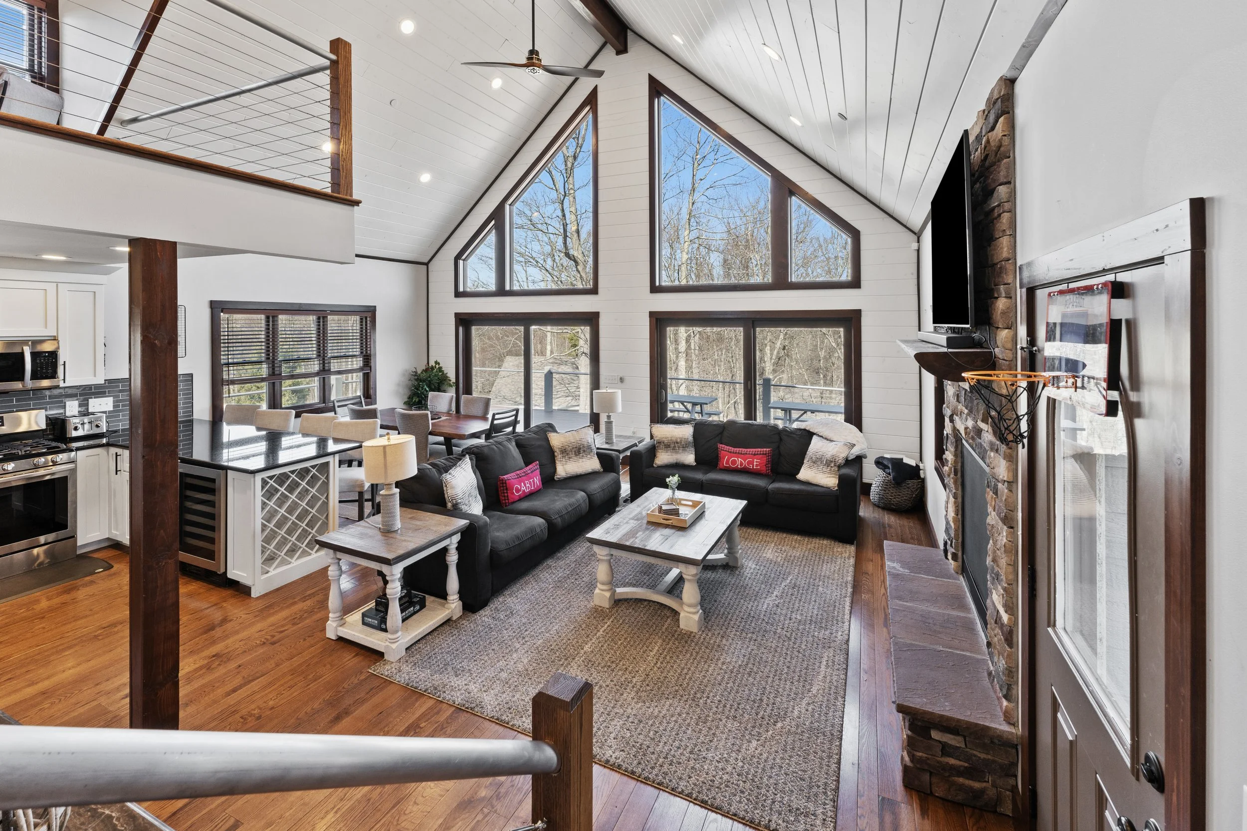 A spacious open living room with high vaulted ceilings, large triangular windows, and wooden accents. There are two black sofas with cushions, a white coffee table, a brick fireplace with a mounted TV, and a wooden floor with a rug. The room also includes a dining area with a table and chairs, and the kitchen with white cabinets and black countertops is partially visible.