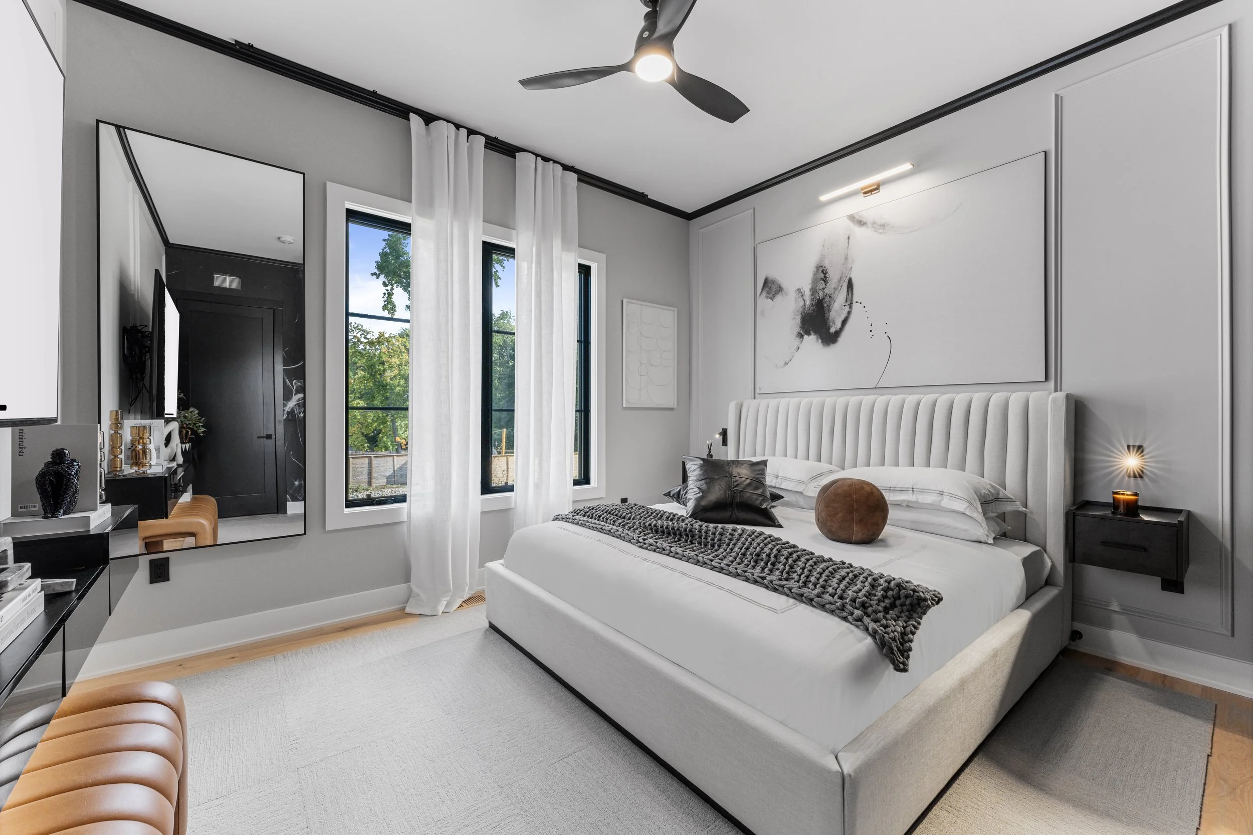 Modern bedroom with a white bed, decorative pillows, black and white artwork above the headboard, black floating nightstand with a candle, a large mirror, white curtains, wide windows, ceiling fan, and minimalist decor.
