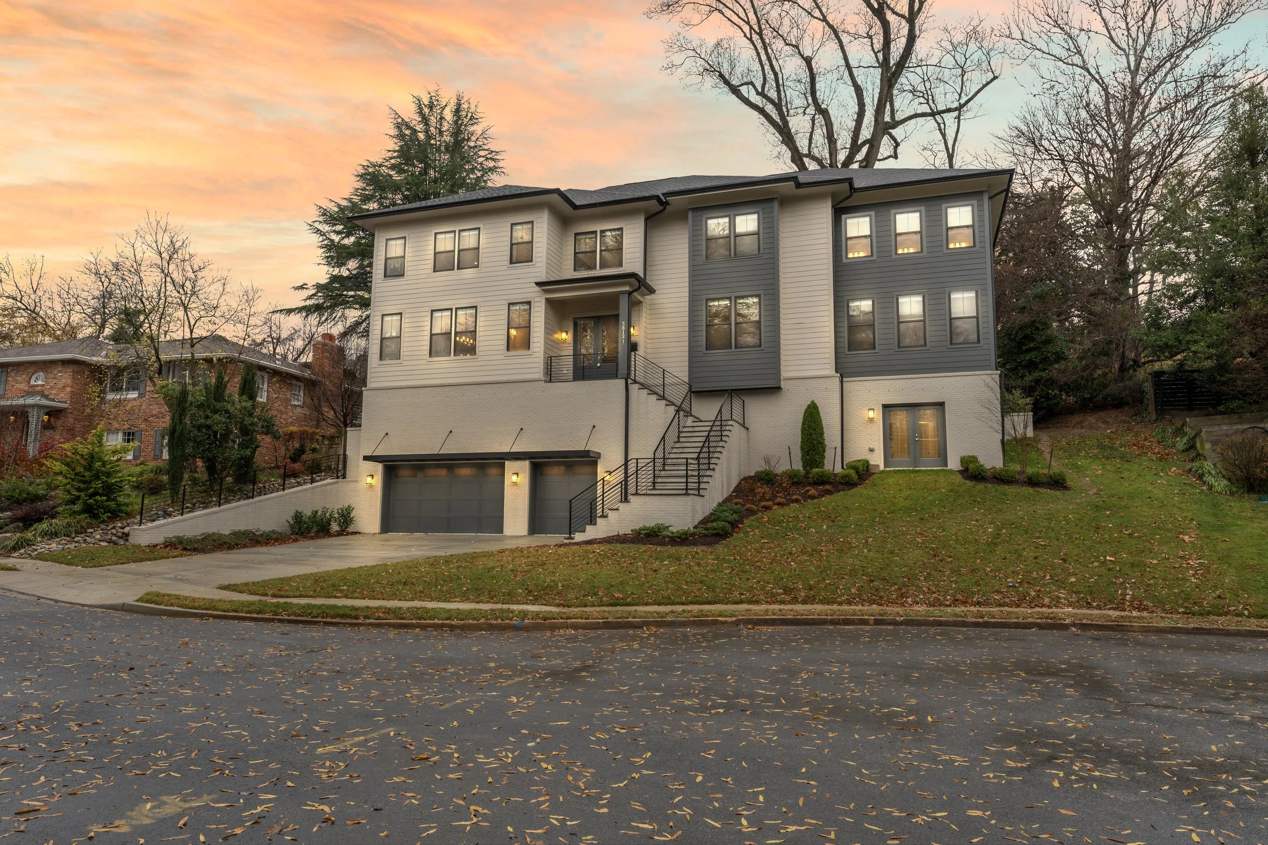 Modern multi-story house with a dark gray and beige exterior, stairs leading to the front door, two garages, and landscaped yard with trees and bushes, during sunset.
