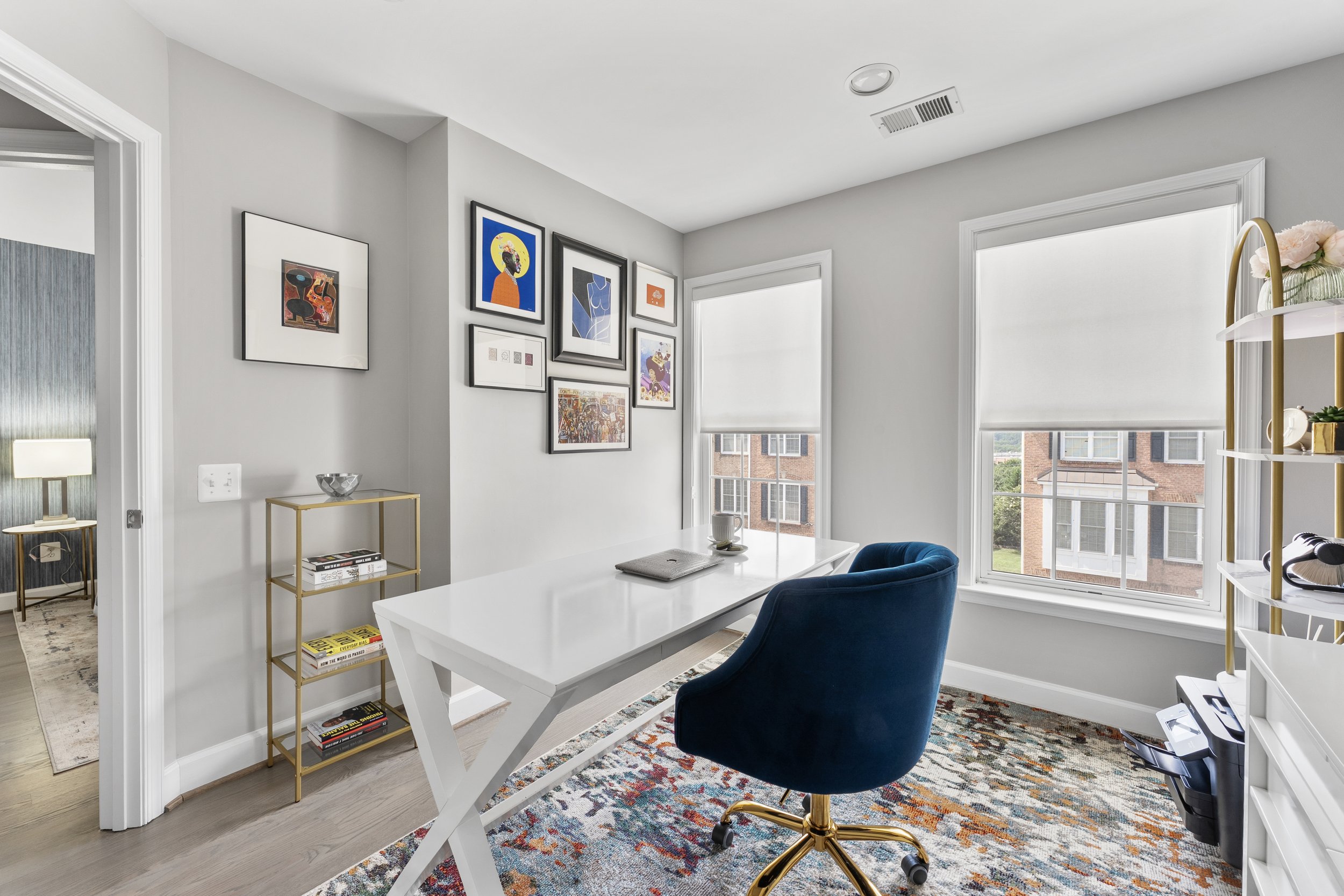 Bright home office with a white desk, a blue velvet swivel chair, a colorful area rug, and a gallery wall of framed artwork on light gray walls. Large windows with white shades let in natural light.