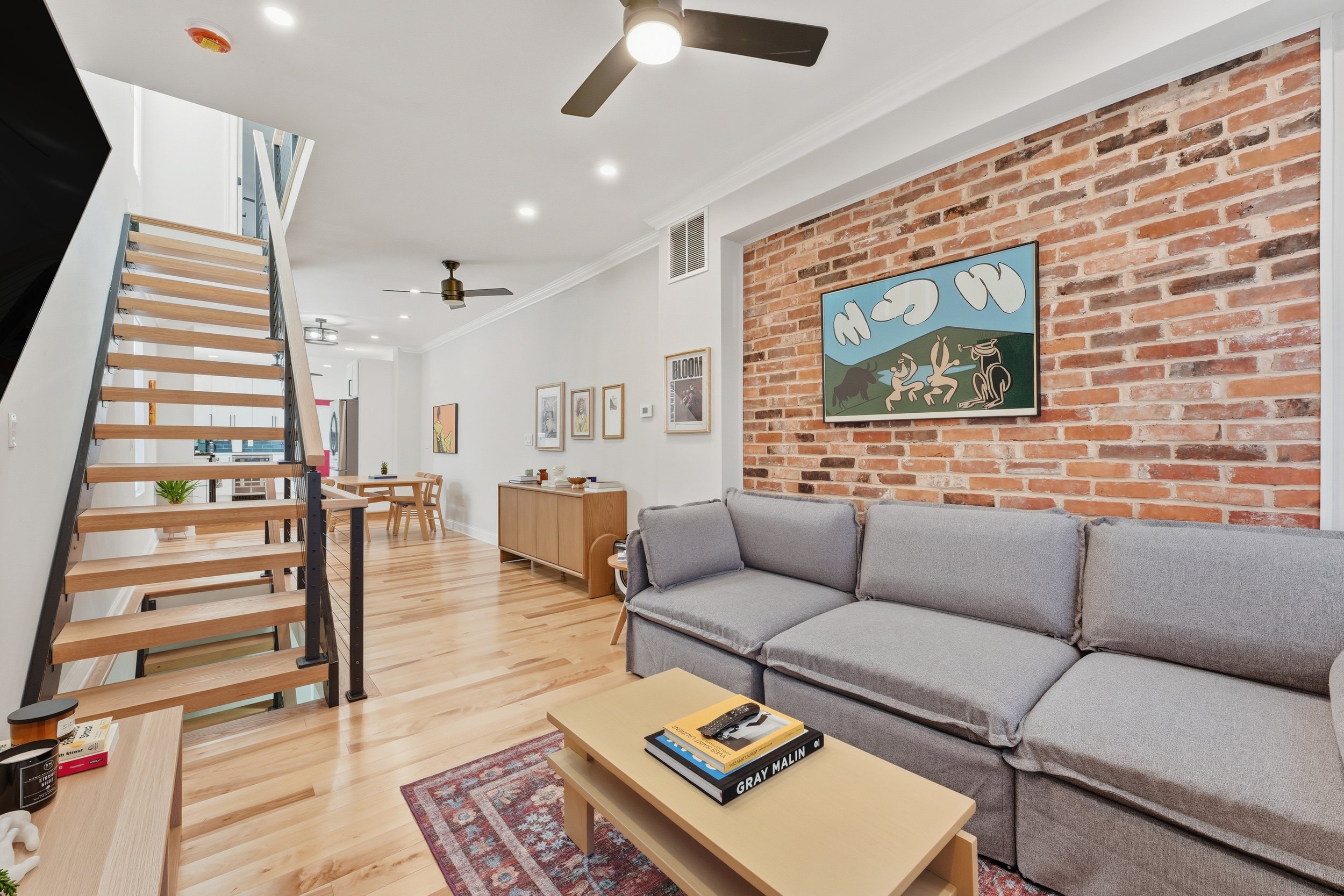 Living room with a gray sectional sofa, brick accent wall with a colorful painting, and a wooden coffee table with books, in a modern open-concept home.