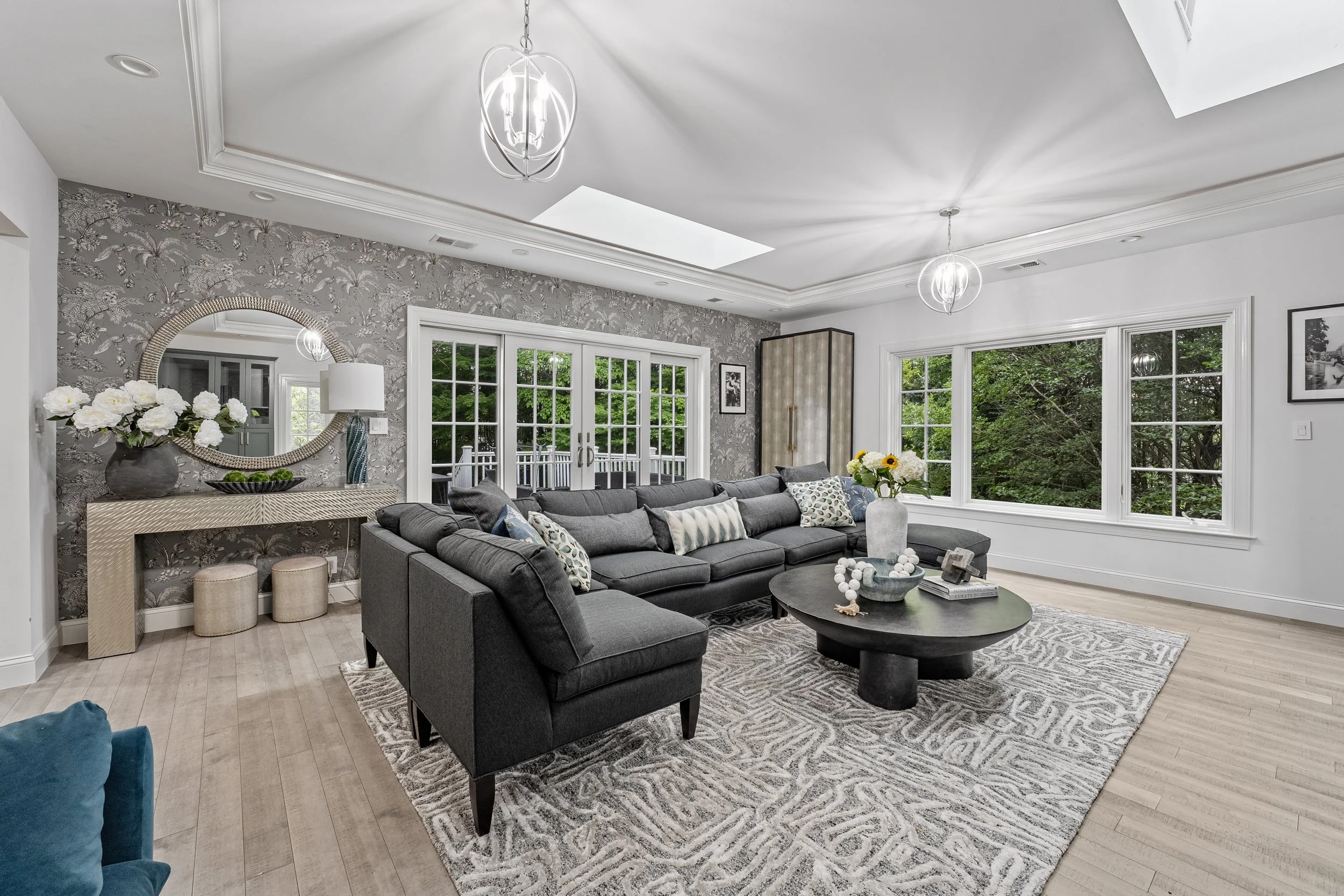 Living room with gray sectional sofa, black coffee table, large window overlooking a green yard, patterned rug, floral wallpaper accent wall, decorative mirror, and modern light fixtures.