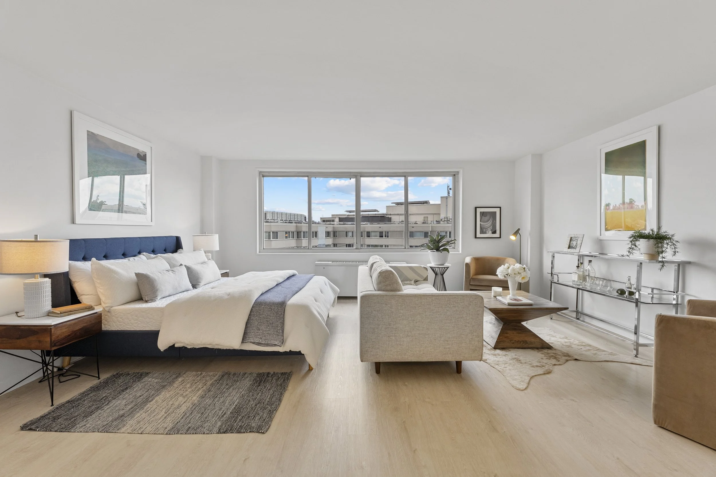 Bright, modern living room with large window, queen bed, sofa, armchairs, wooden and metallic furniture, artwork, and decorative plants.