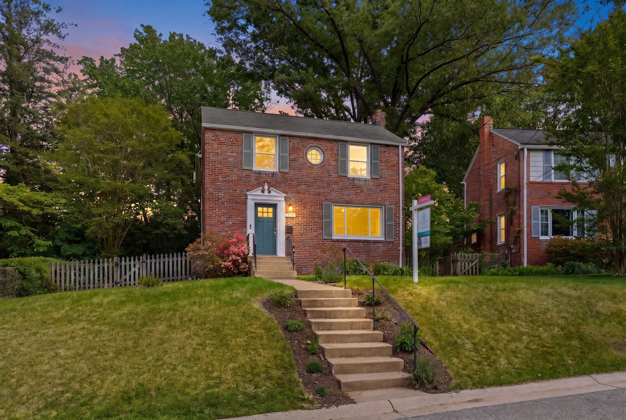 Red brick colonial home at twilight with warmly lit windows, a sloped front lawn and a pink and blue sunset sky