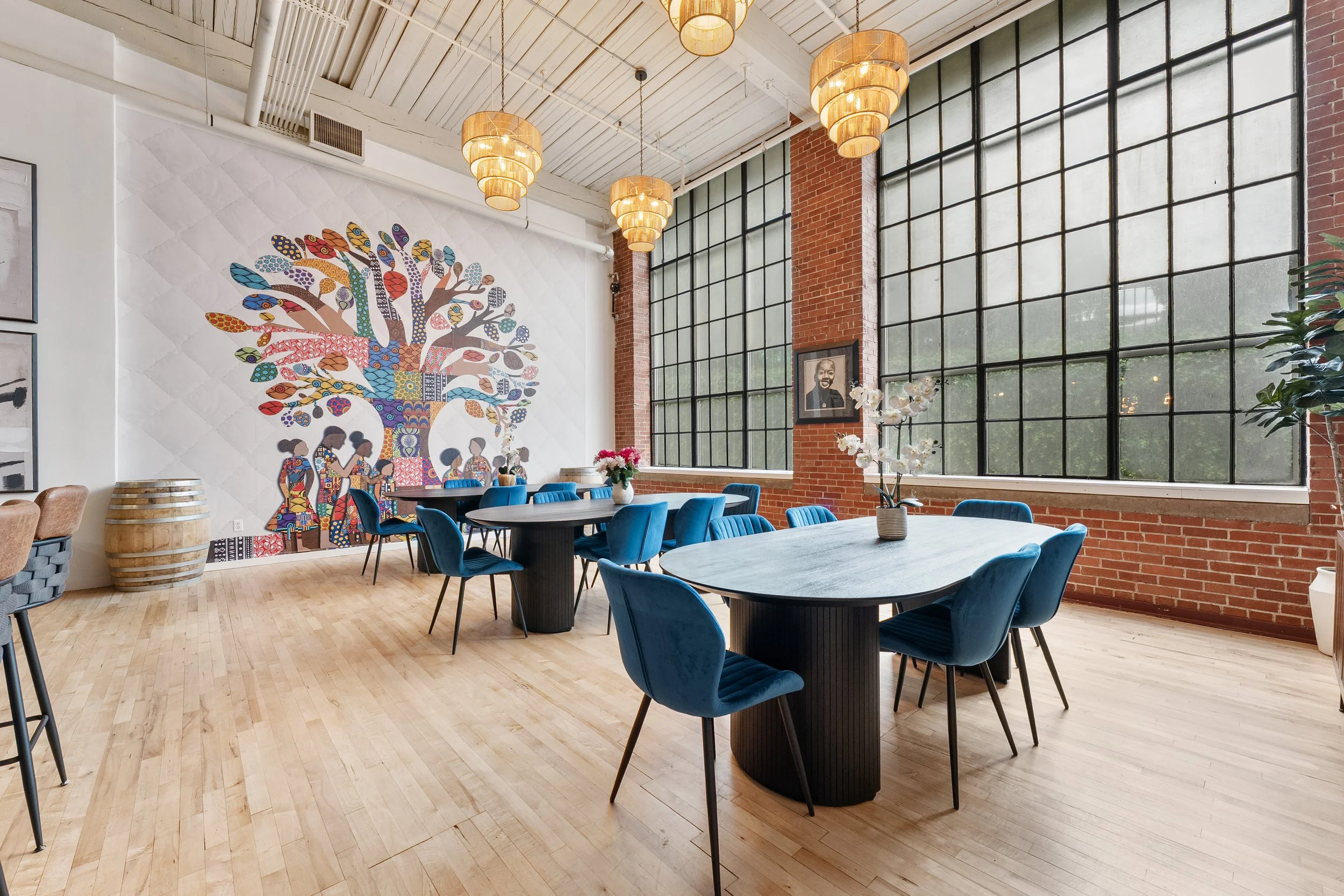 Interior of a modern restaurant or cafe with wooden floors, large windows, blue chairs around black tables, and colorful artwork of a tree on the wall.