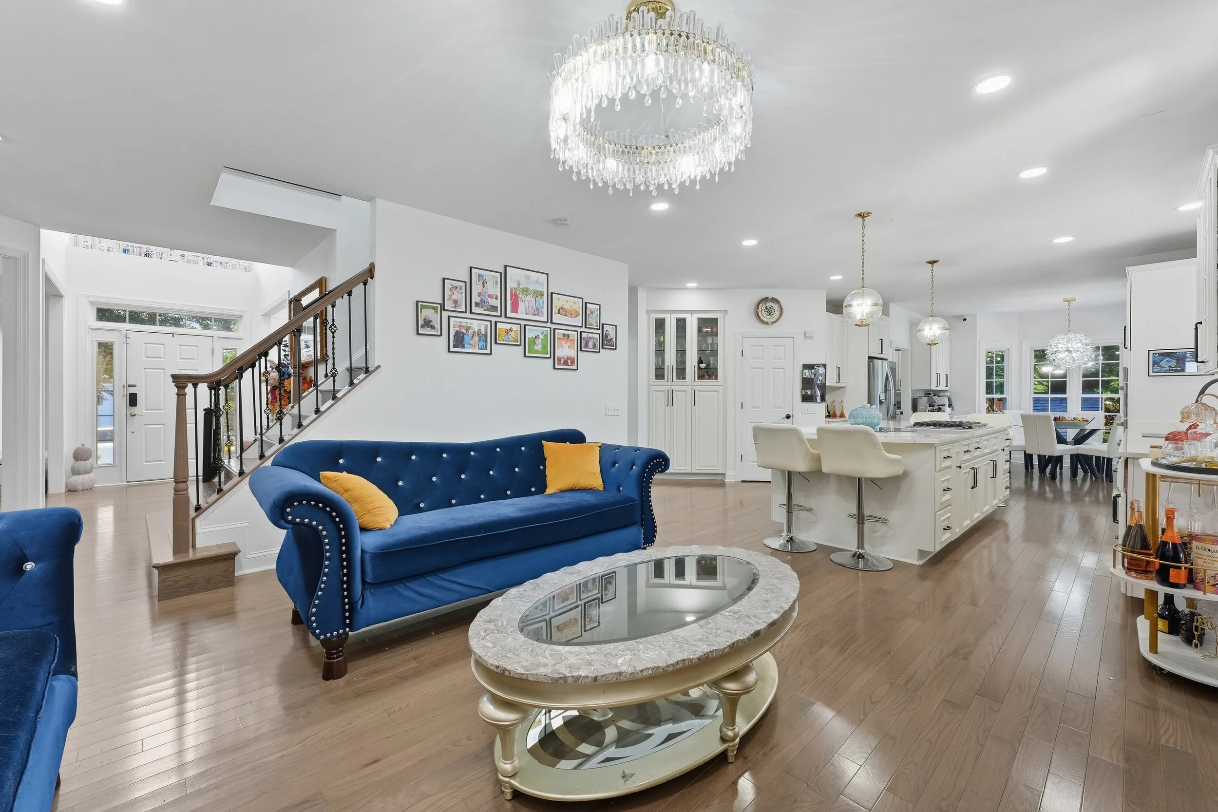 Open living room with a blue velvet sofa and crystal chandelier, opening to a white kitchen beyond.