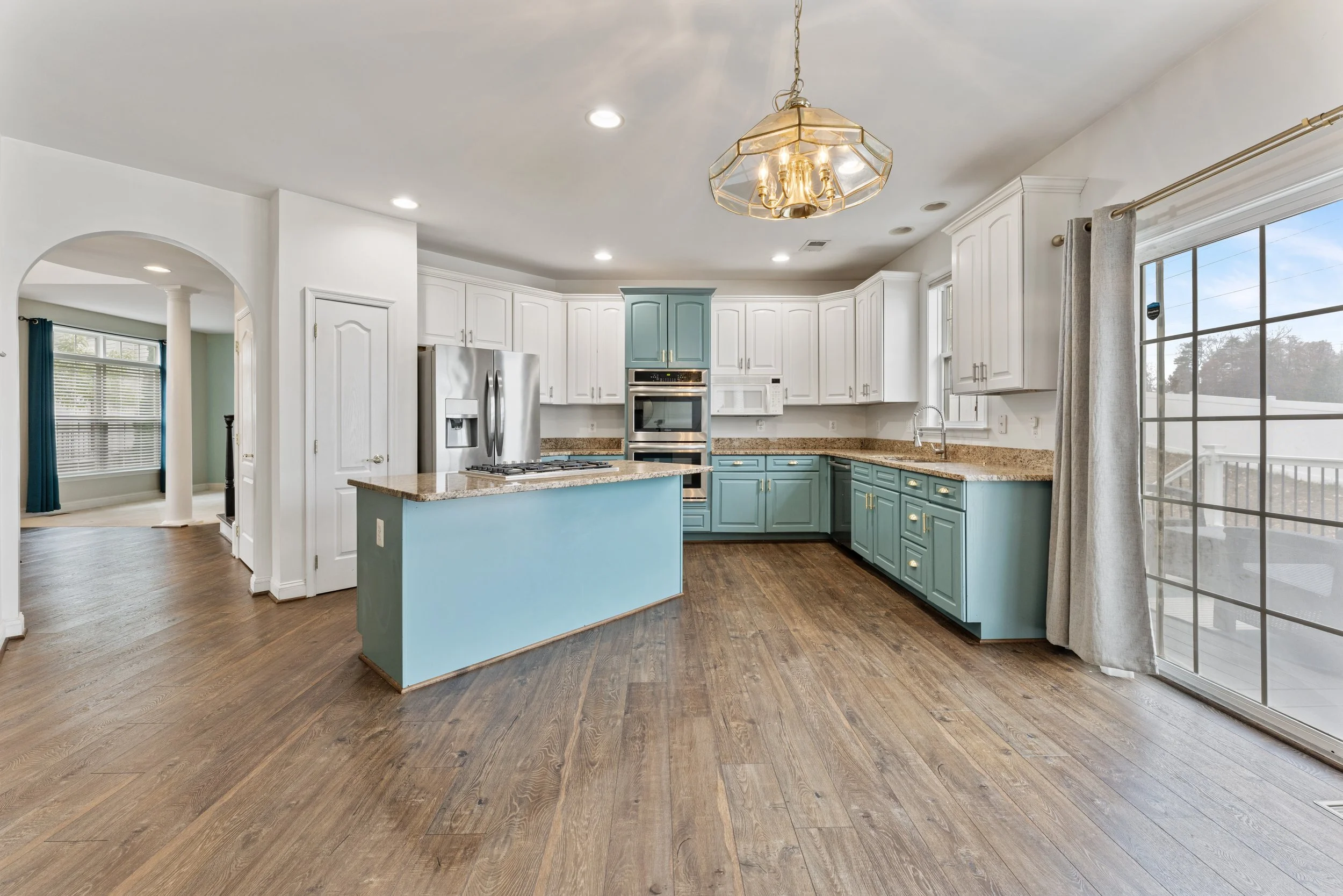 Kitchen with white and turquoise cabinets, stainless steel refrigerator, granite countertops, hardwood flooring, and large window with sliding door.
