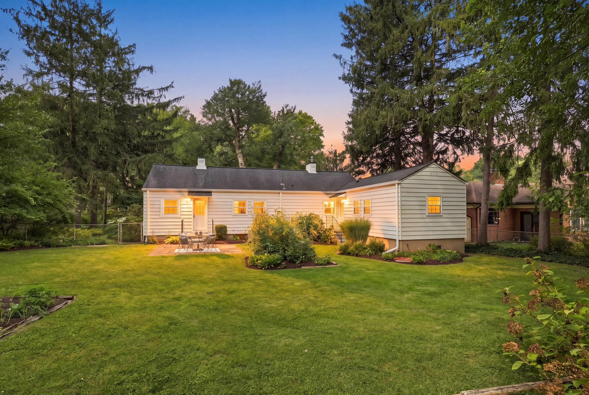 Rear exterior of a white ranch home at dusk with warmly lit windows and a landscaped lawn