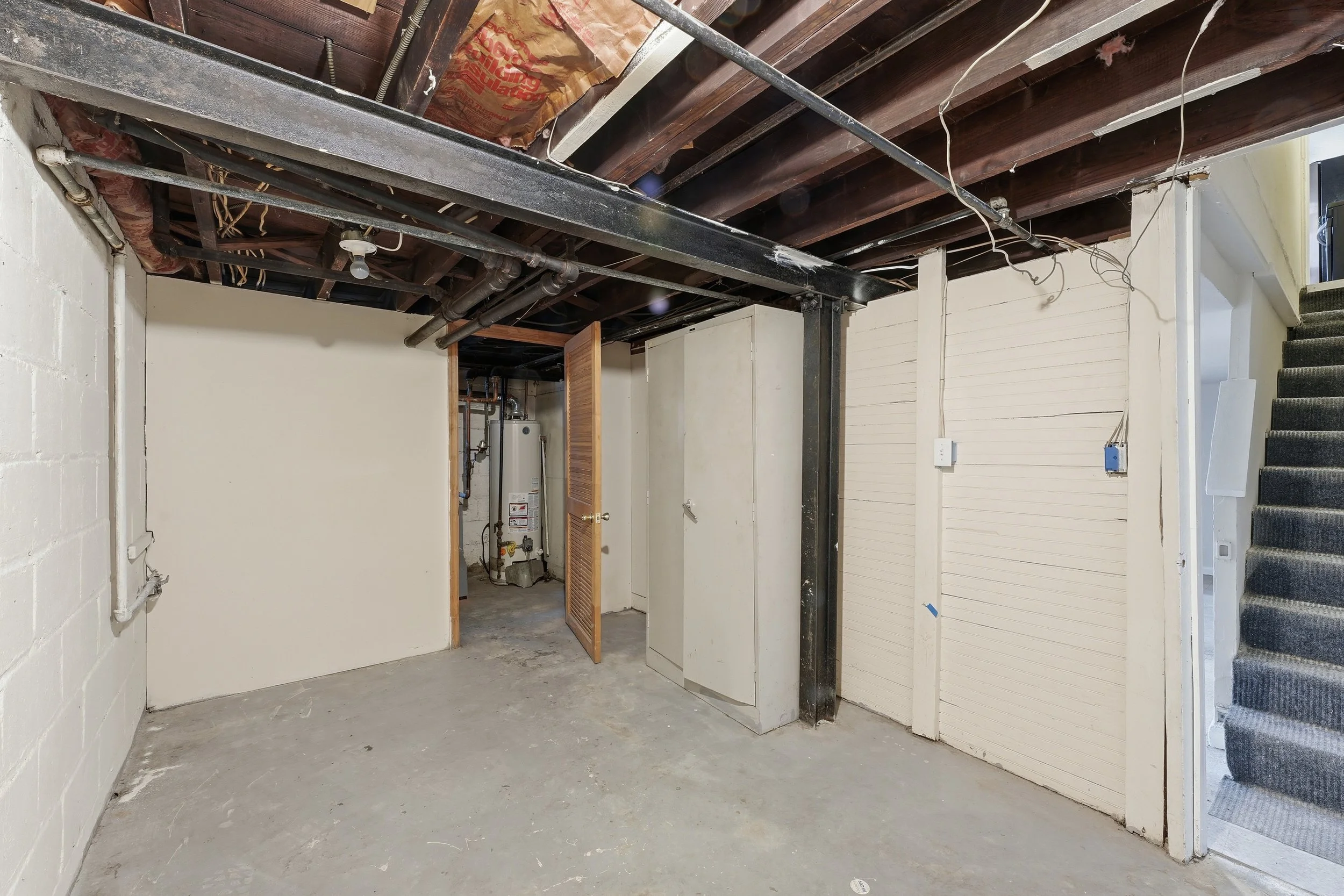 Unfinished basement with exposed joists, pipes and insulation overhead, a bare-bulb fixture, painted block walls, a concrete floor, a carpeted stairway on the right and a water heater through a doorway.