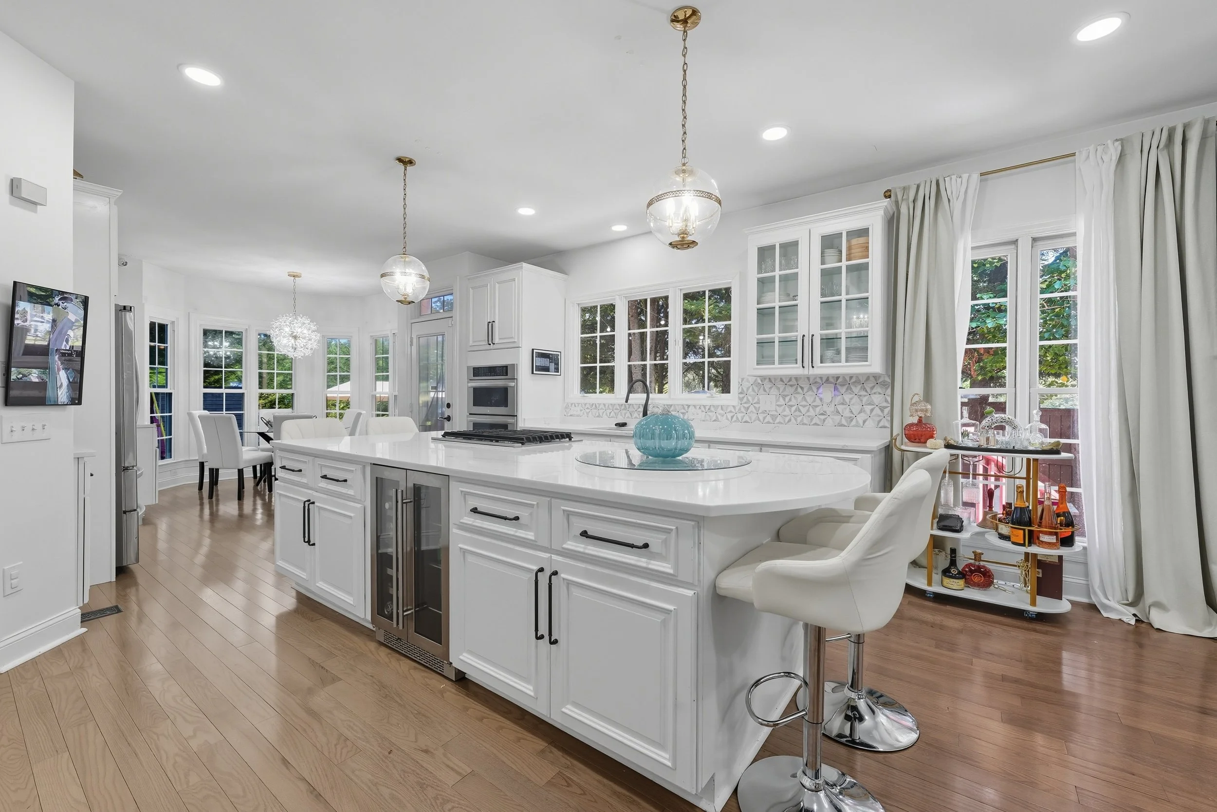 White kitchen with a large quartz island, brass pendant lights, and windows onto trees, with a dining area beyond.