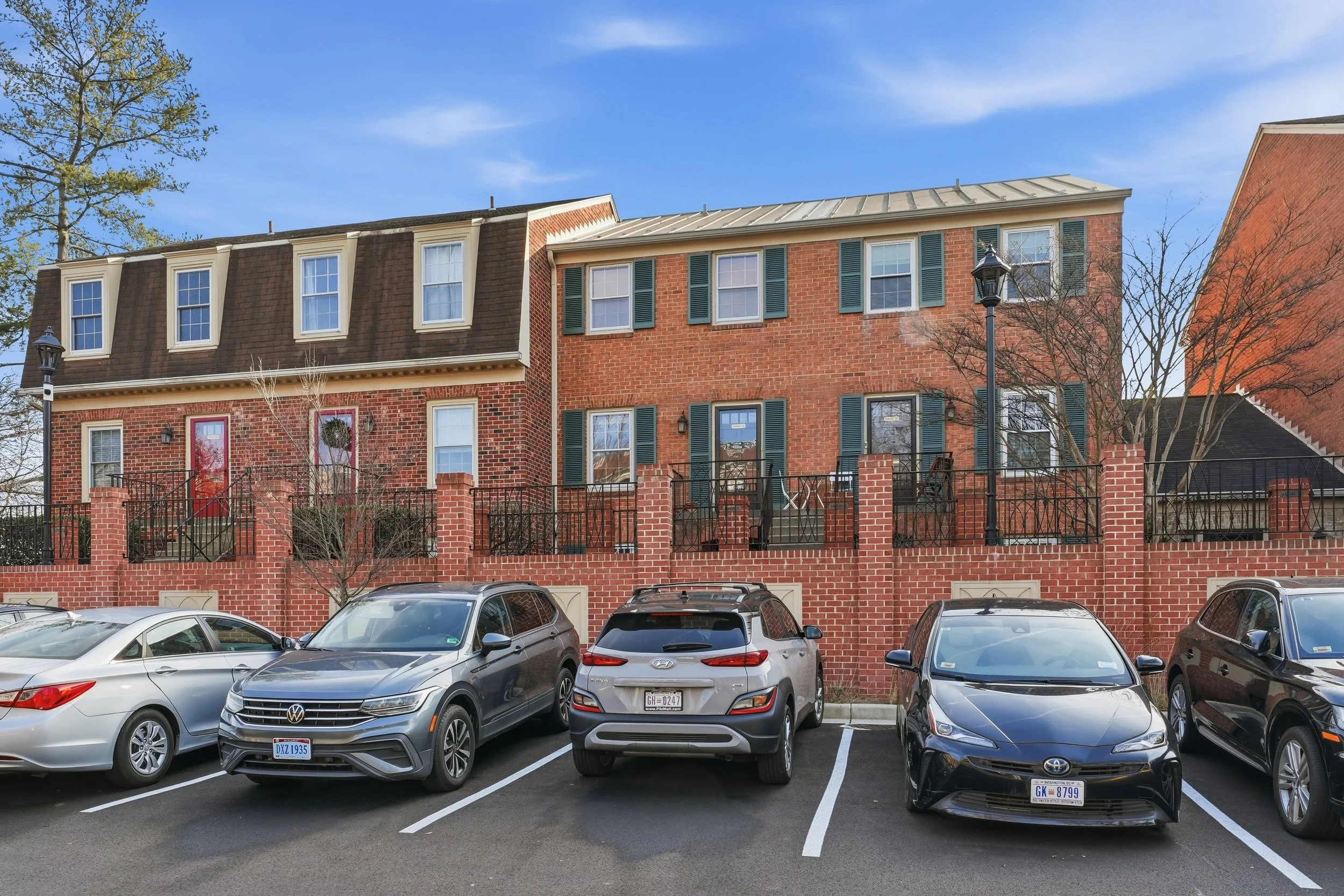 Row of brick townhouses in Northwest DC with green shutters, black iron railings and brick pillars, bare trees, a wreath on one of two red front doors, and cars parked in the lot out front.