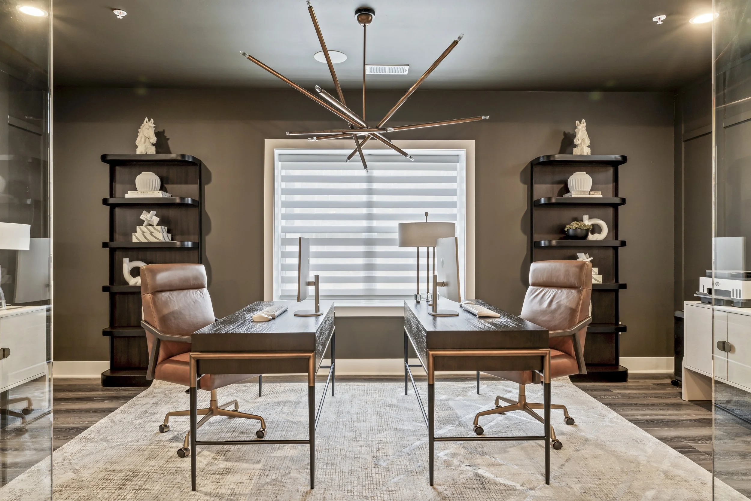 Modern office with two desks, leather chairs, black bookshelves, decorative vases, sculptures, a window with blinds, and a contemporary chandelier.