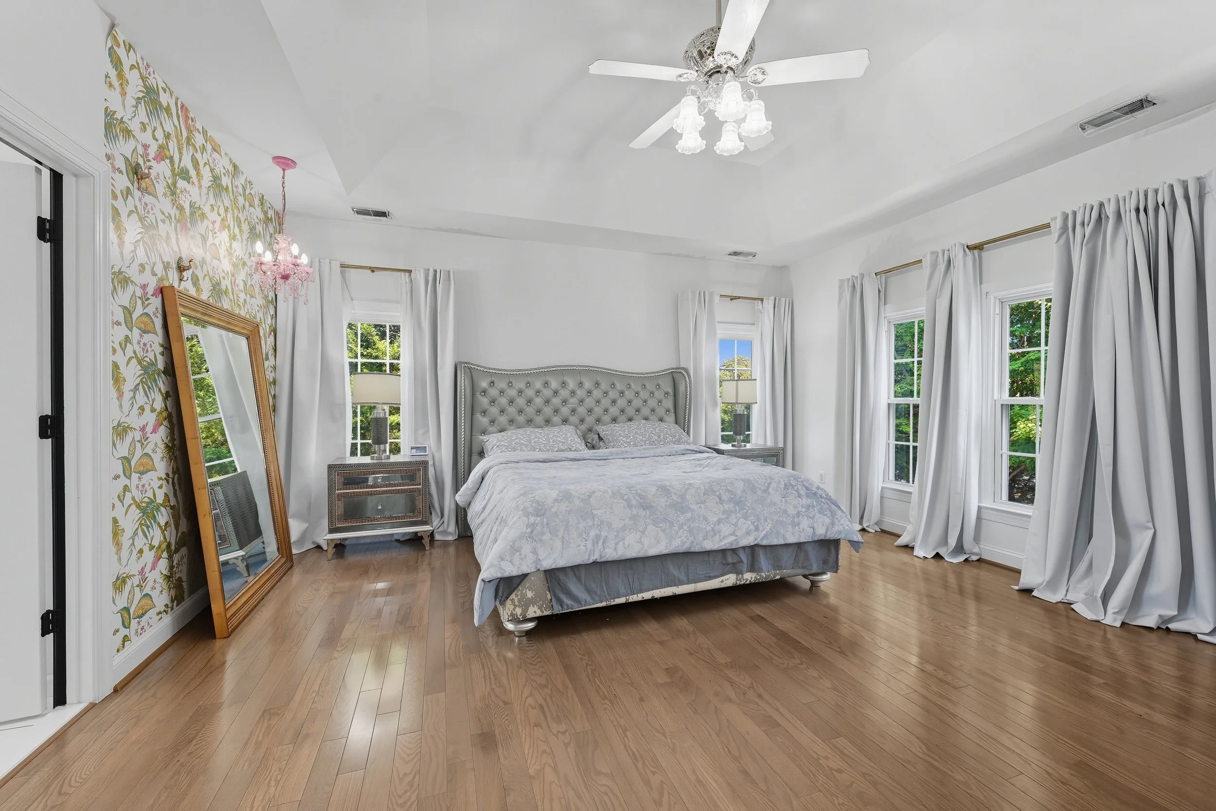Primary bedroom with a floral accent wall, upholstered bed, leaning floor mirror, and windows on two sides over warm oak floors.
