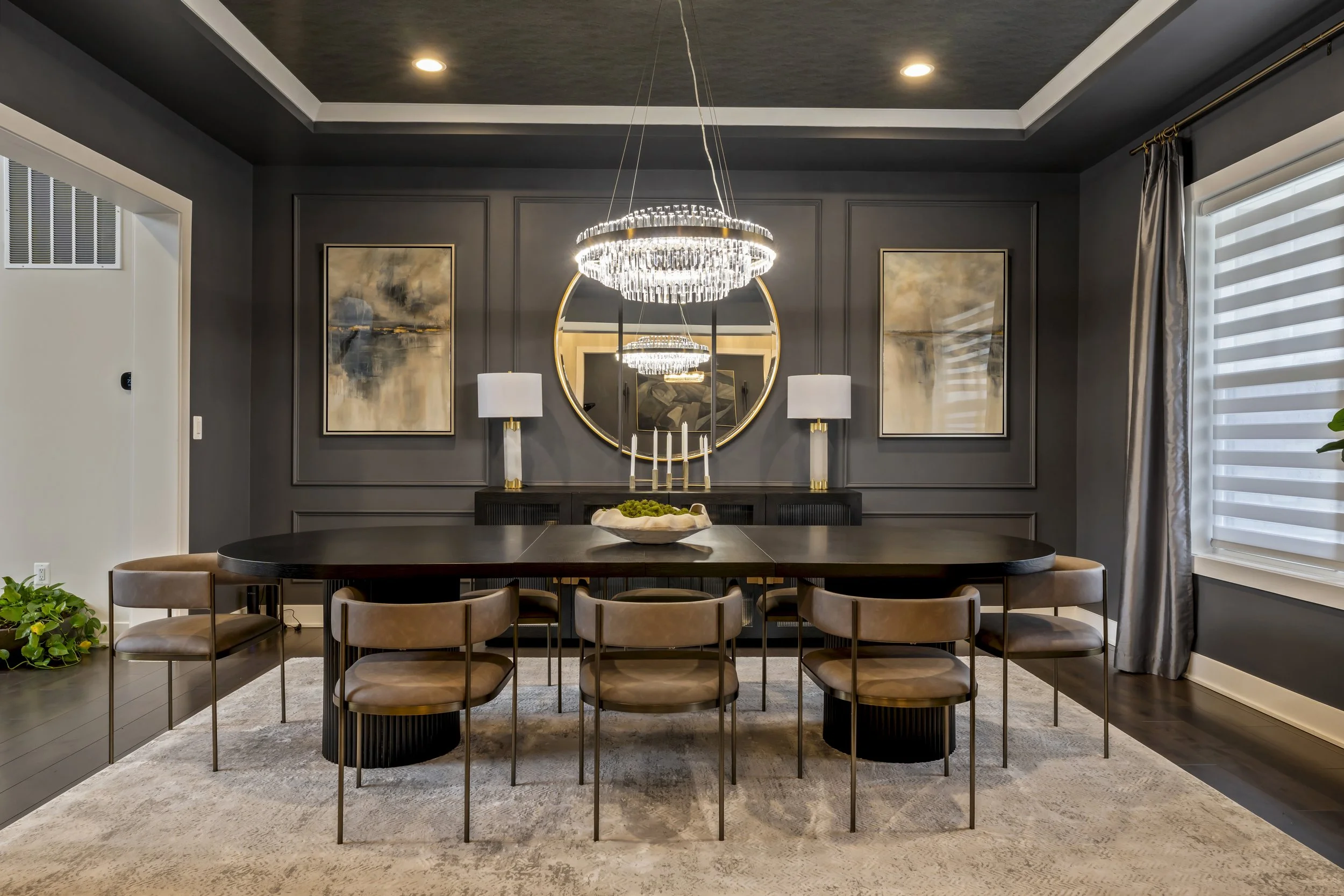 Modern dining room with dark gray walls, black wooden dining table, beige chairs, large mirror, two white lamps, abstract paintings, chandelier, window with blinds, curtain, and a beige rug.