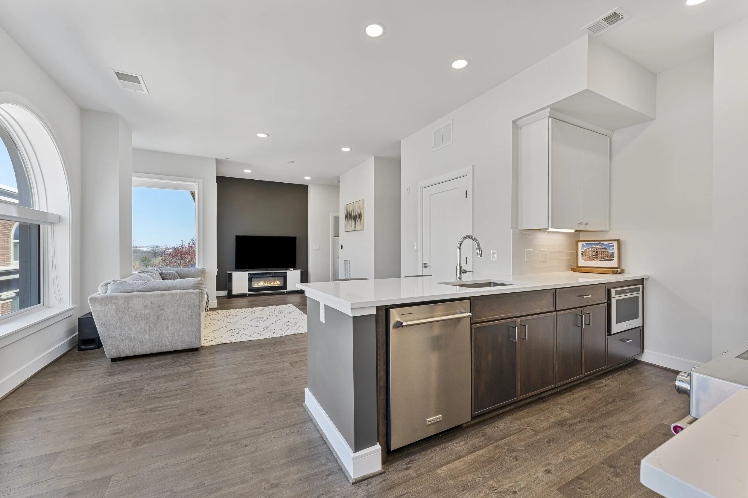 Open-concept living room and kitchen with white walls, large windows, a gray sofa, a television, a fireplace, and hardwood floors.