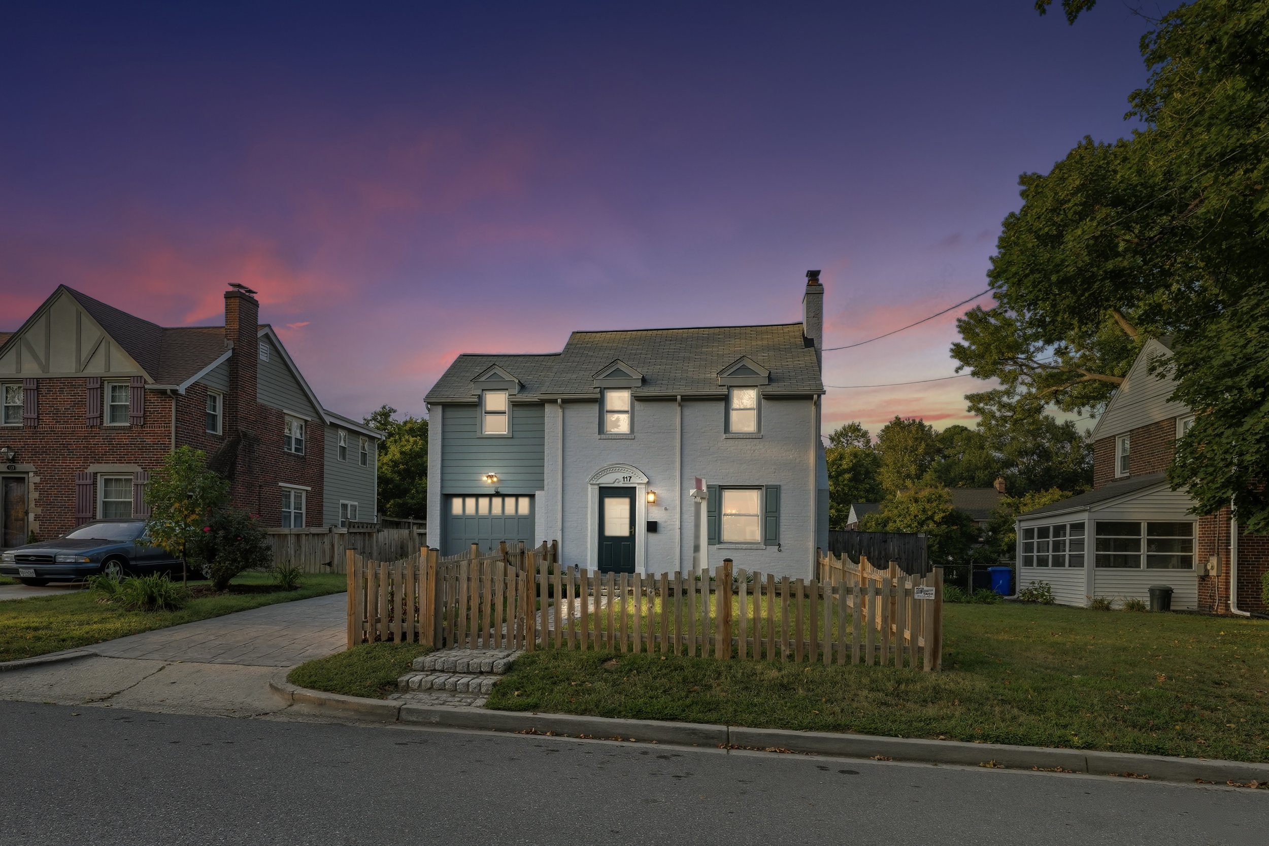 A two-story white house with a black door, front porch light on, small fenced yard, surrounded by neighboring houses, trees, and a colorful sunset sky.