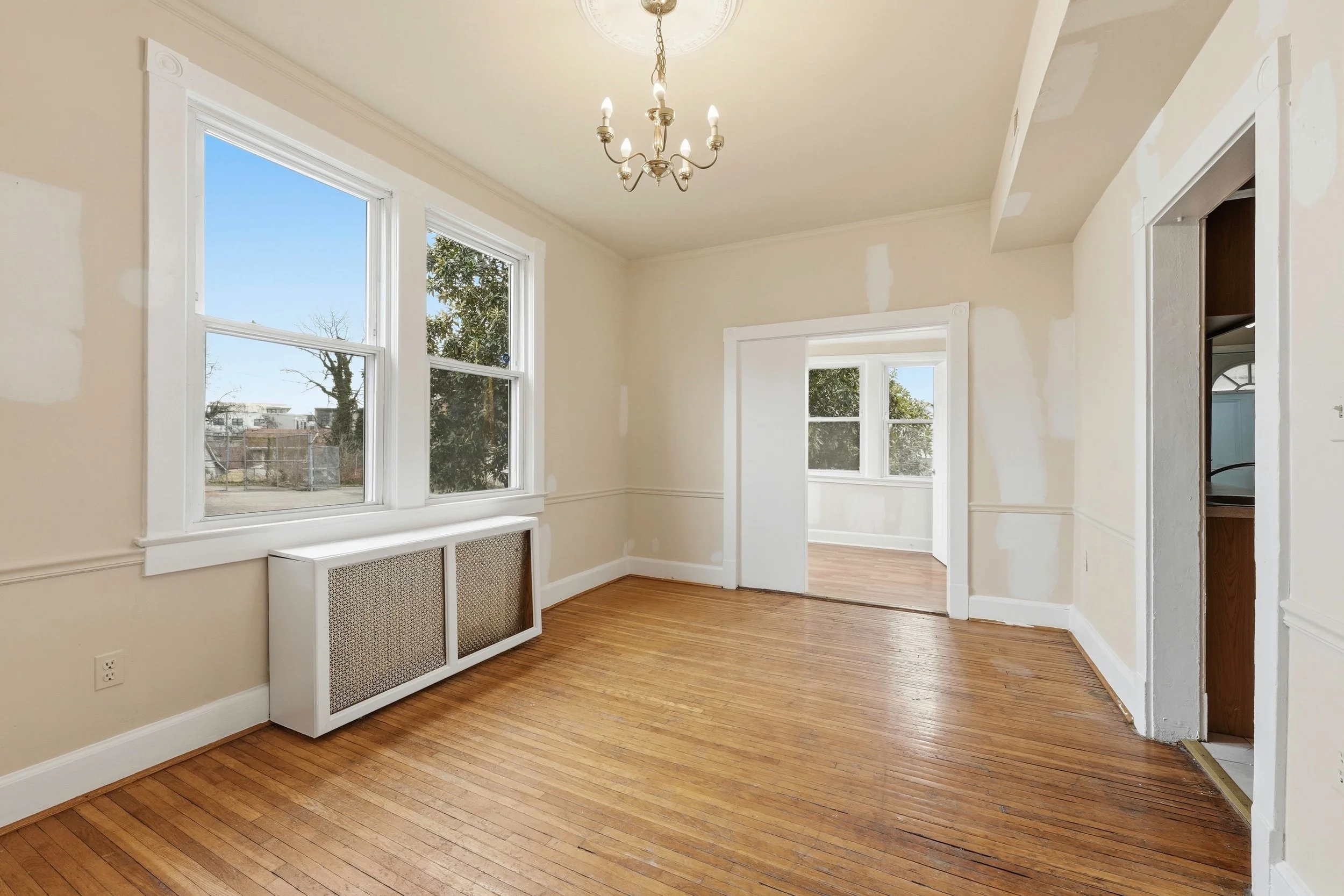 Empty dining room with hardwood floors, a small brass chandelier, a white radiator cover under two tall windows showing trees outside, and a doorway to a sunroom.