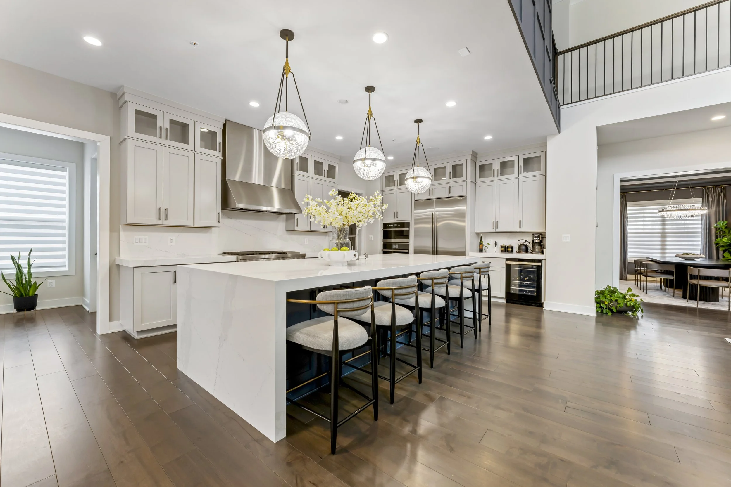 Modern kitchen with white cabinetry, stainless steel appliances, and a large island with seating. There are pendant lights hanging above the island, and a sliding glass door with blinds in the background.