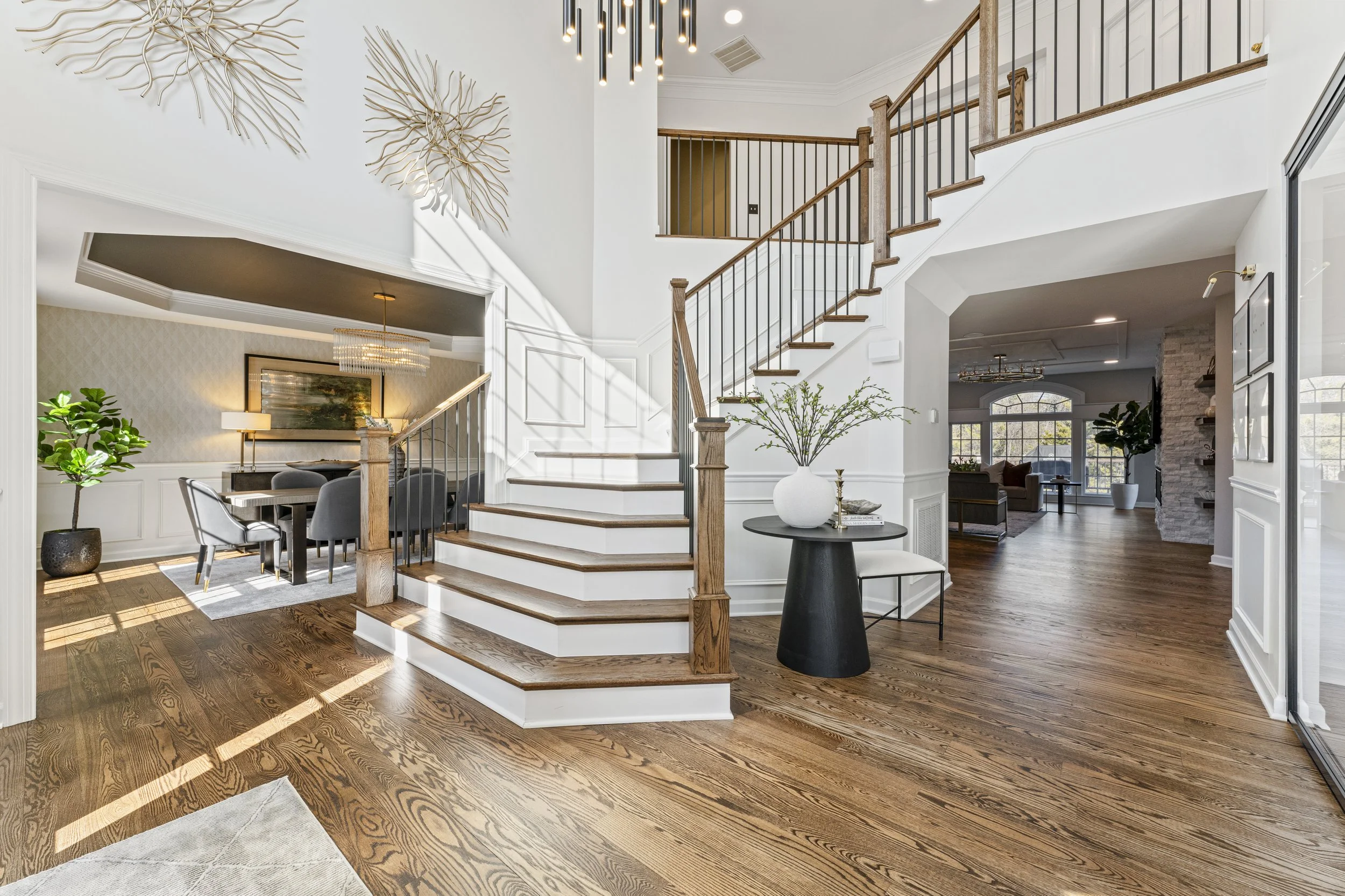 Modern living space with hardwood floors, white walls, staircase with wooden railing, and decorative wall art. Contains an entryway with a small table and plant, and views into various room areas.