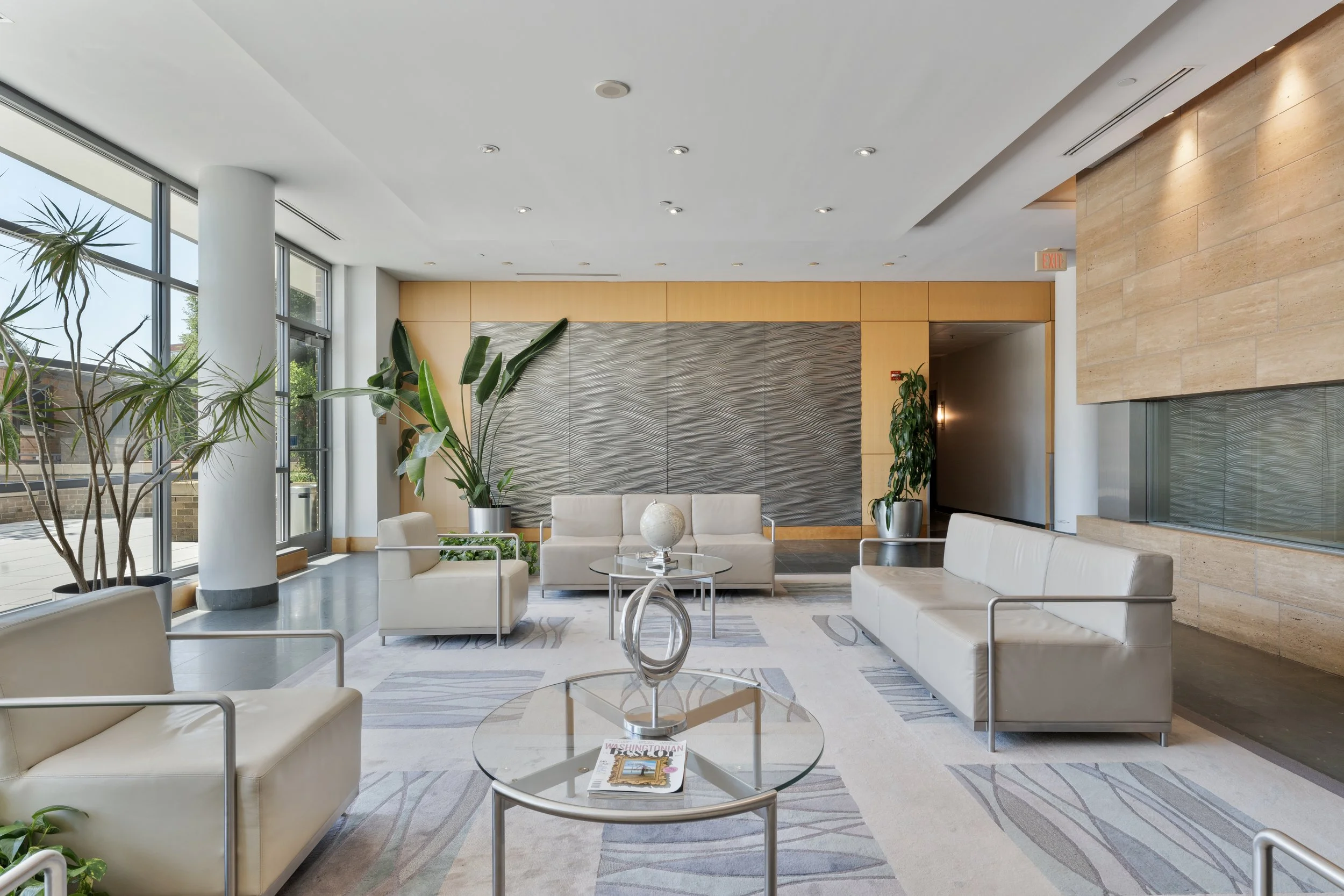 Modern hotel lobby with white sofas, glass tables, large plants, and a textured wall decor, illuminated by natural light from floor-to-ceiling windows.