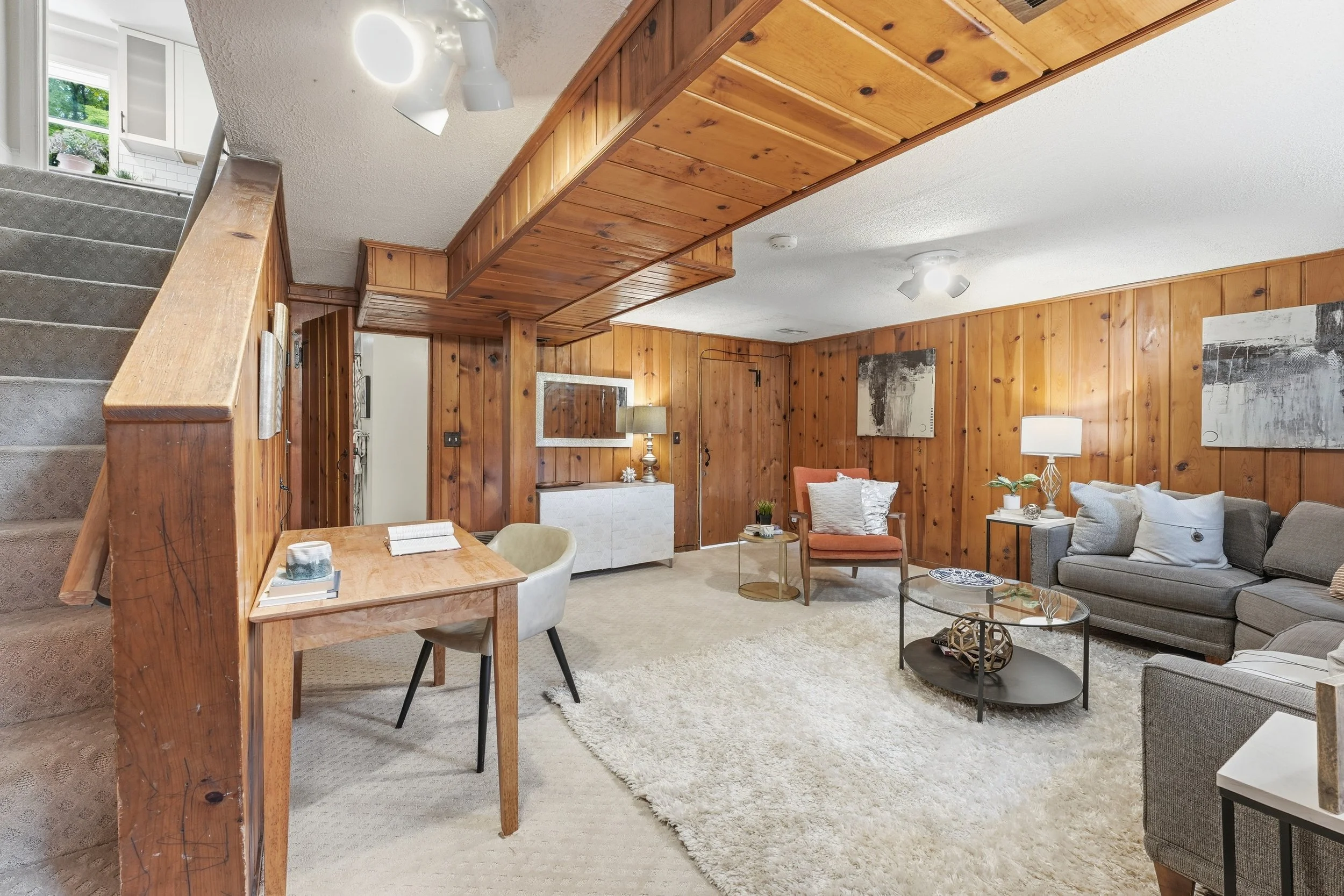 Rec room with knotty-pine paneled walls and a beamed ceiling, a gray sectional and a shag rug, a small desk beside a carpeted staircase, and a window at the top of the stairs.