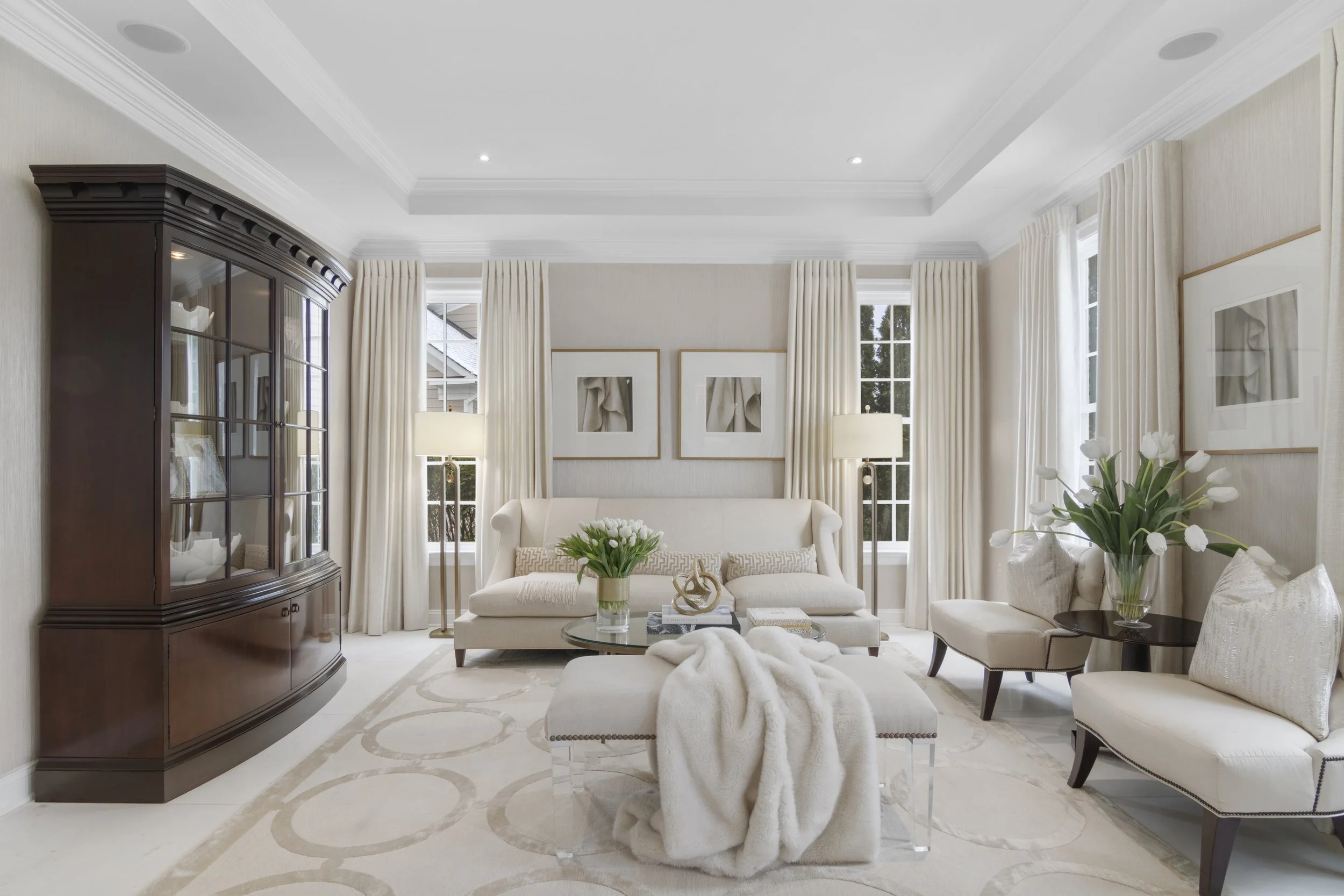 Elegant living room with white sofas, floral arrangements, beige curtains, framed artwork, a dark wood display cabinet, and large windows.