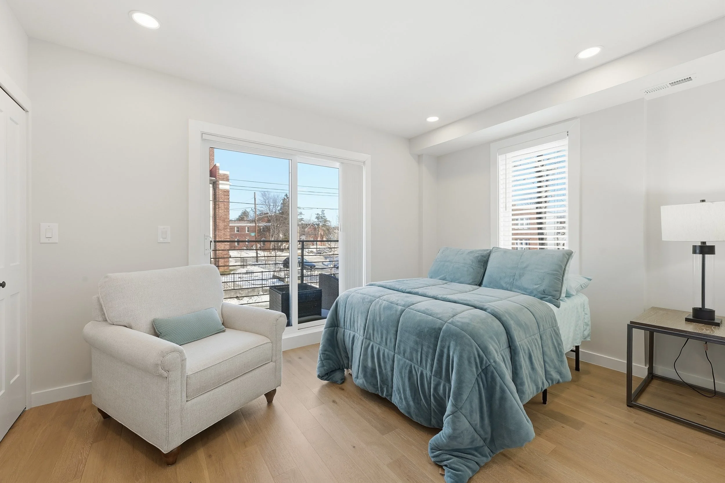 Bright bedroom with a teal comforter, a white armchair and a table lamp, and a sliding glass door to a balcony overlooking snow-covered ground and brick buildings on a sunny winter afternoon.