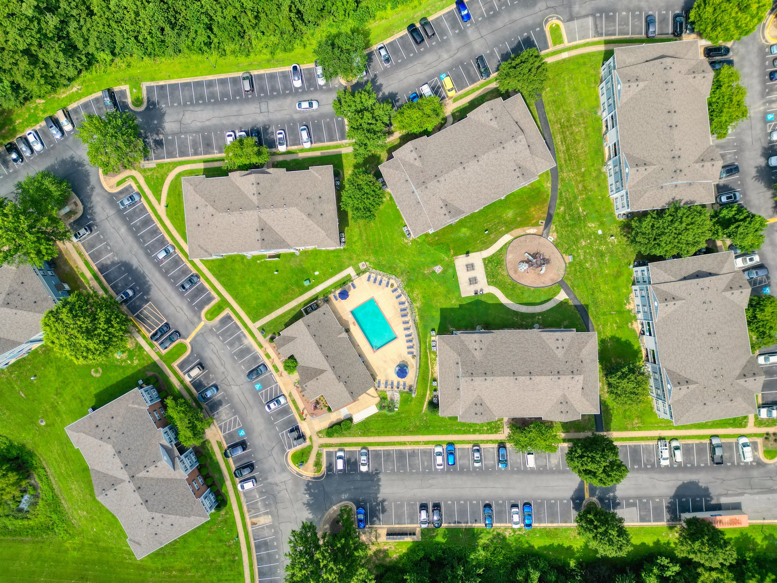 Aerial view of an apartment complex with multiple buildings, green lawns, trees, a swimming pool, a playground, and parking lots with cars.