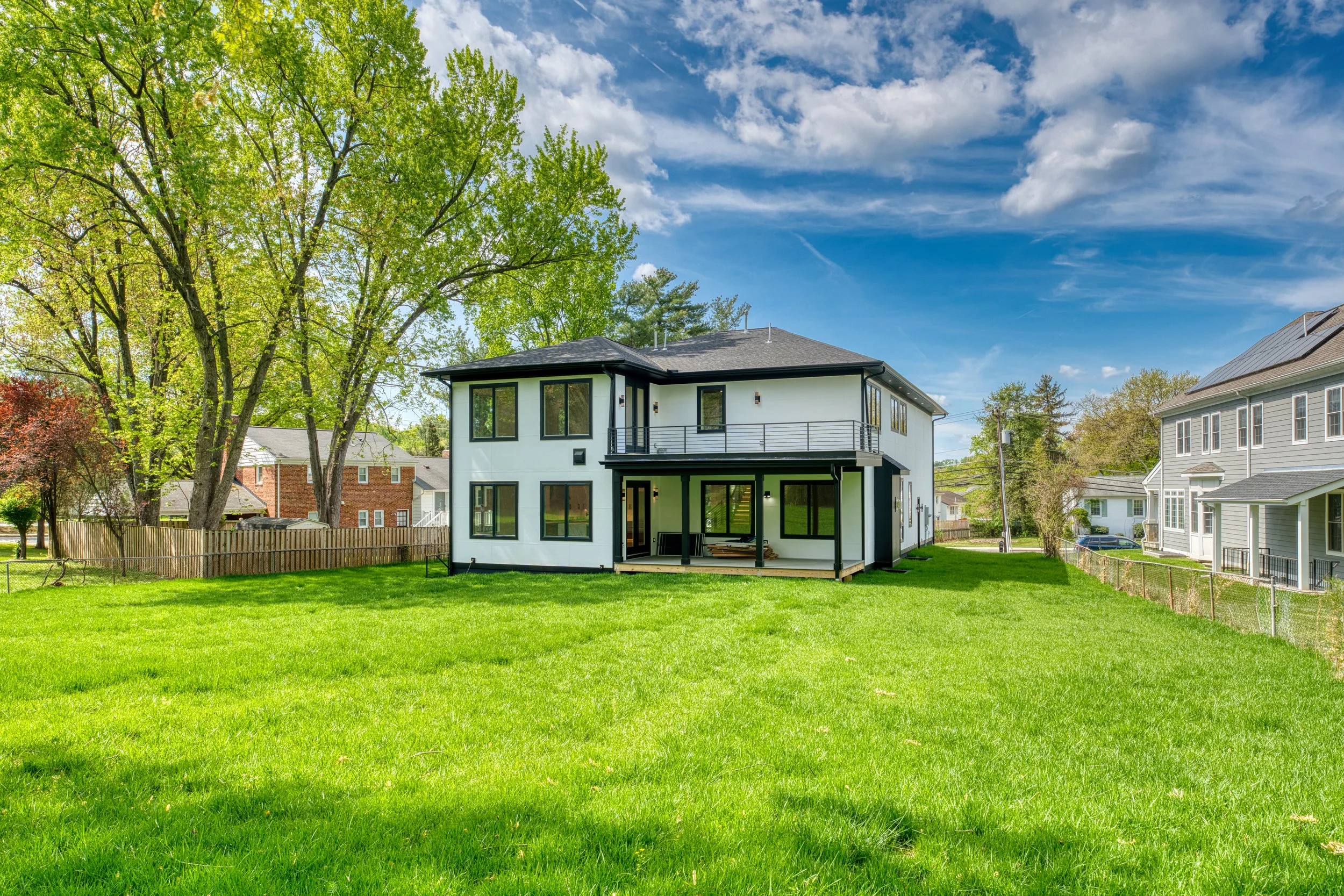 A modern two-story house with black trim, large windows, and a balcony, situated on a grassy lawn with trees and neighboring houses in the background, under a partly cloudy sky.