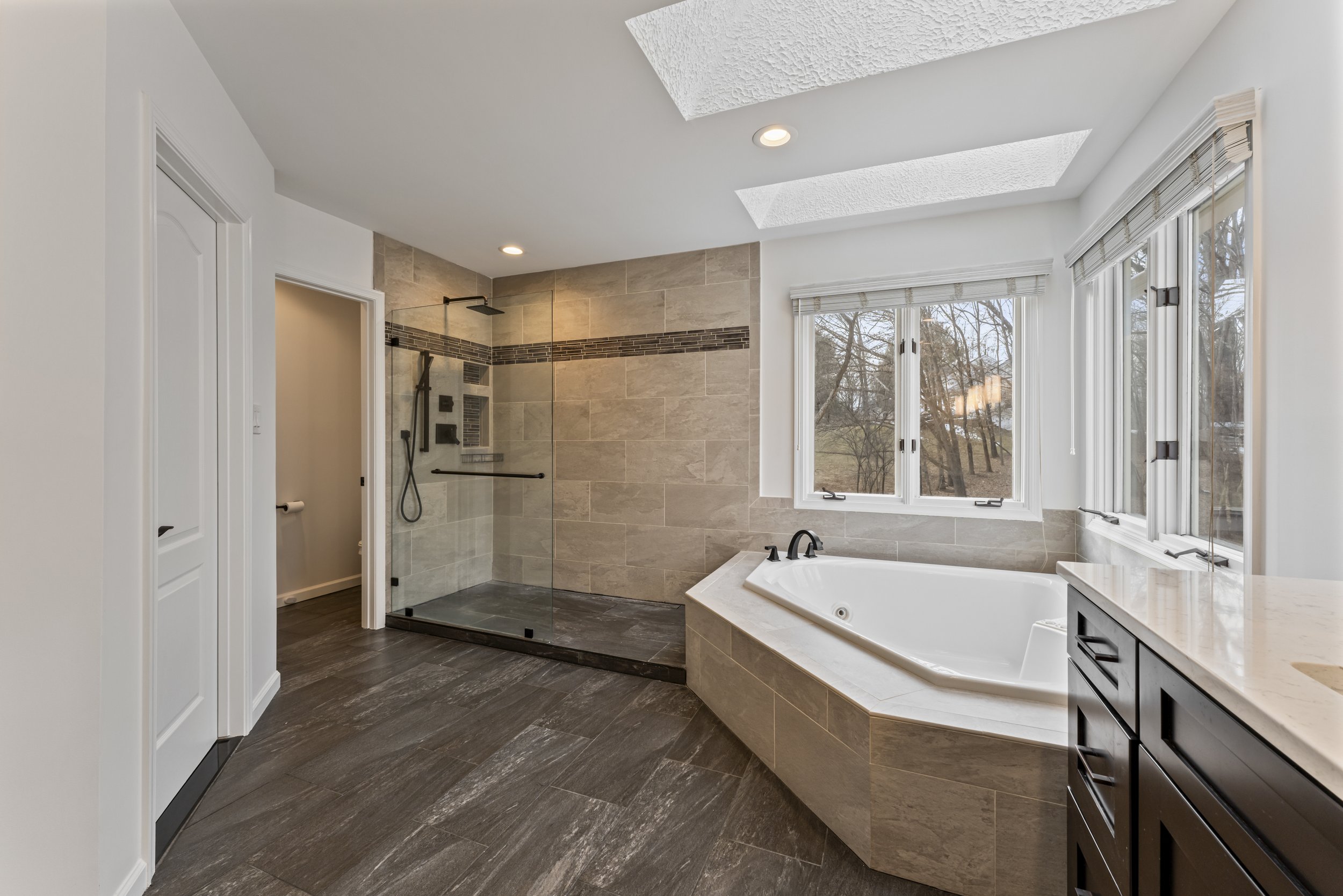 Modern bathroom with glass-enclosed shower, bathtub, large window, dark wood vanity, beige tile walls, and wood-look tile flooring.