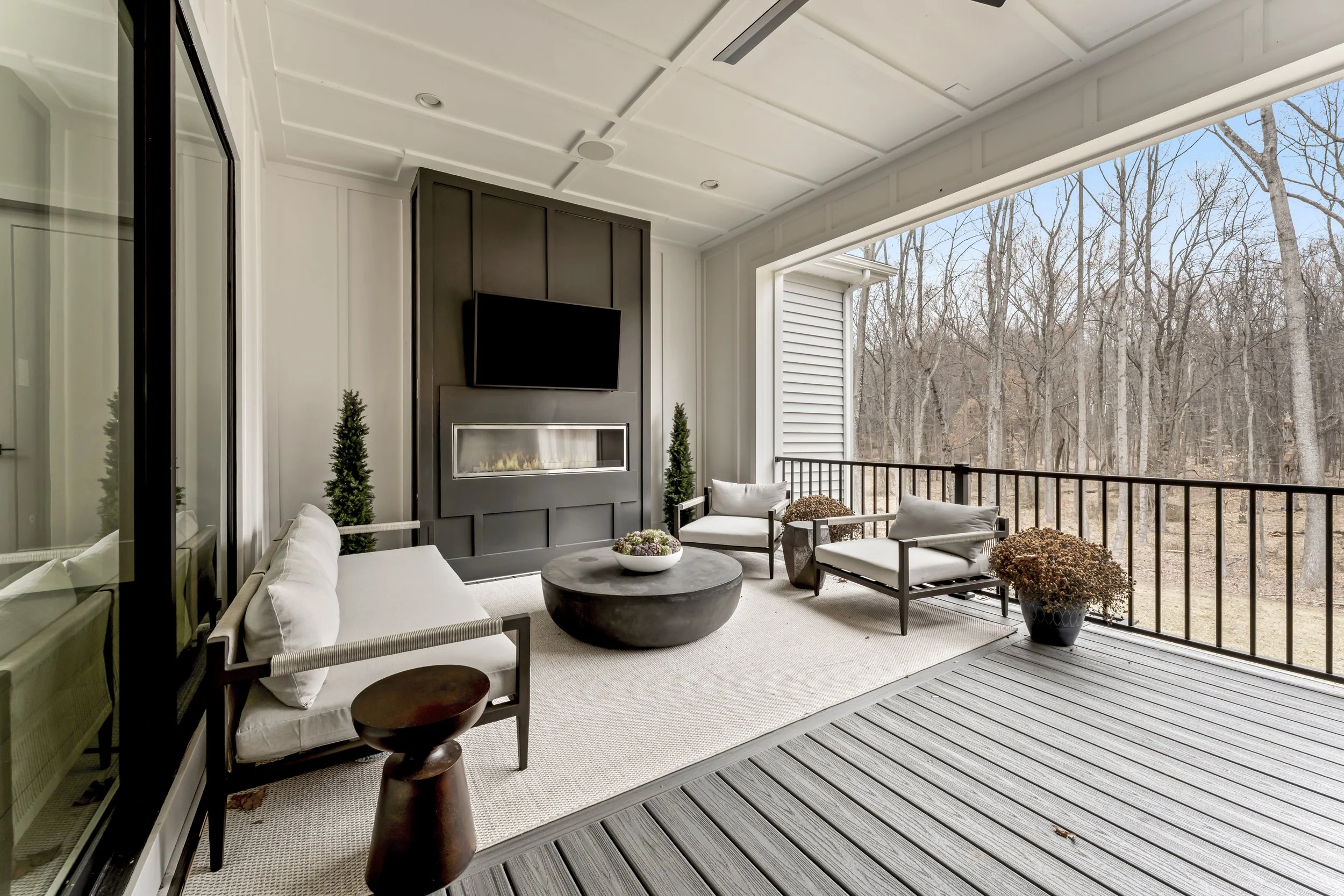 Outdoor balcony with seating area, black fireplace, wall-mounted TV, and view of leafless trees.