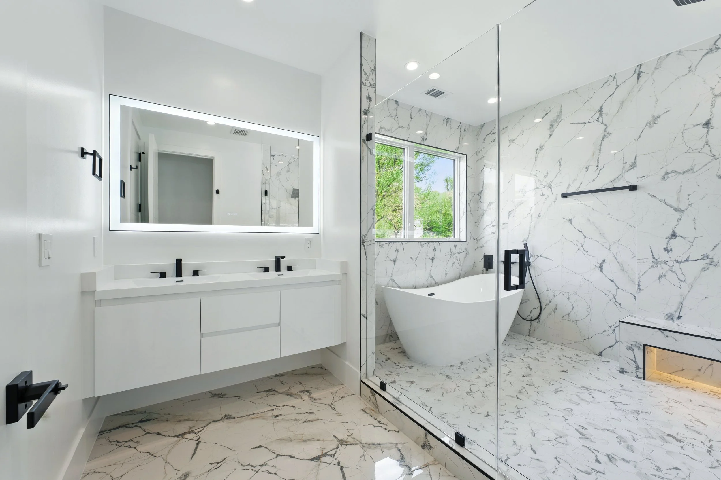 Modern bathroom with white marble walls and floor, black fixtures, a double vanity with backlit mirror, a bathtub near a window, and a glass-enclosed shower.