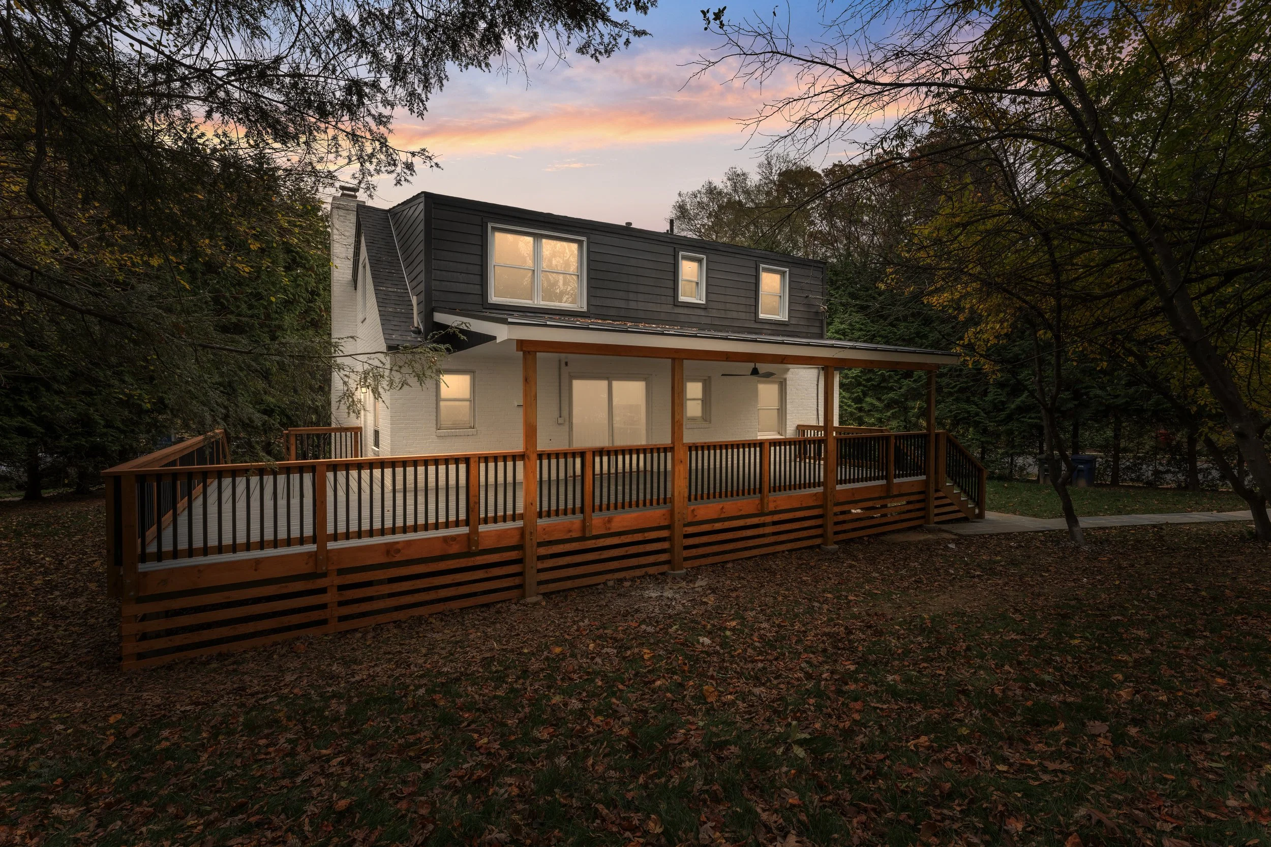 A two-story house with a white brick exterior and a dark-colored roof, featuring a large wooden deck with railings, set in a wooded area during sunset.