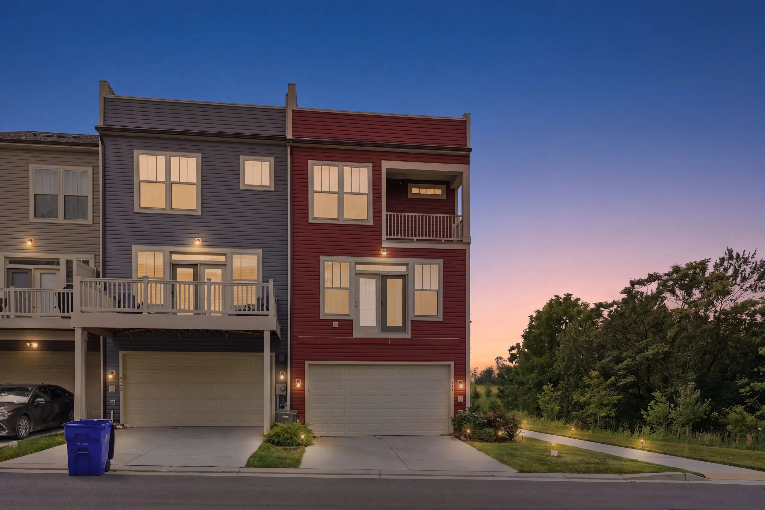 Rear of a row of multi-level townhomes at twilight, with lit windows, upper-level balconies and two-car garages on the ground floor.