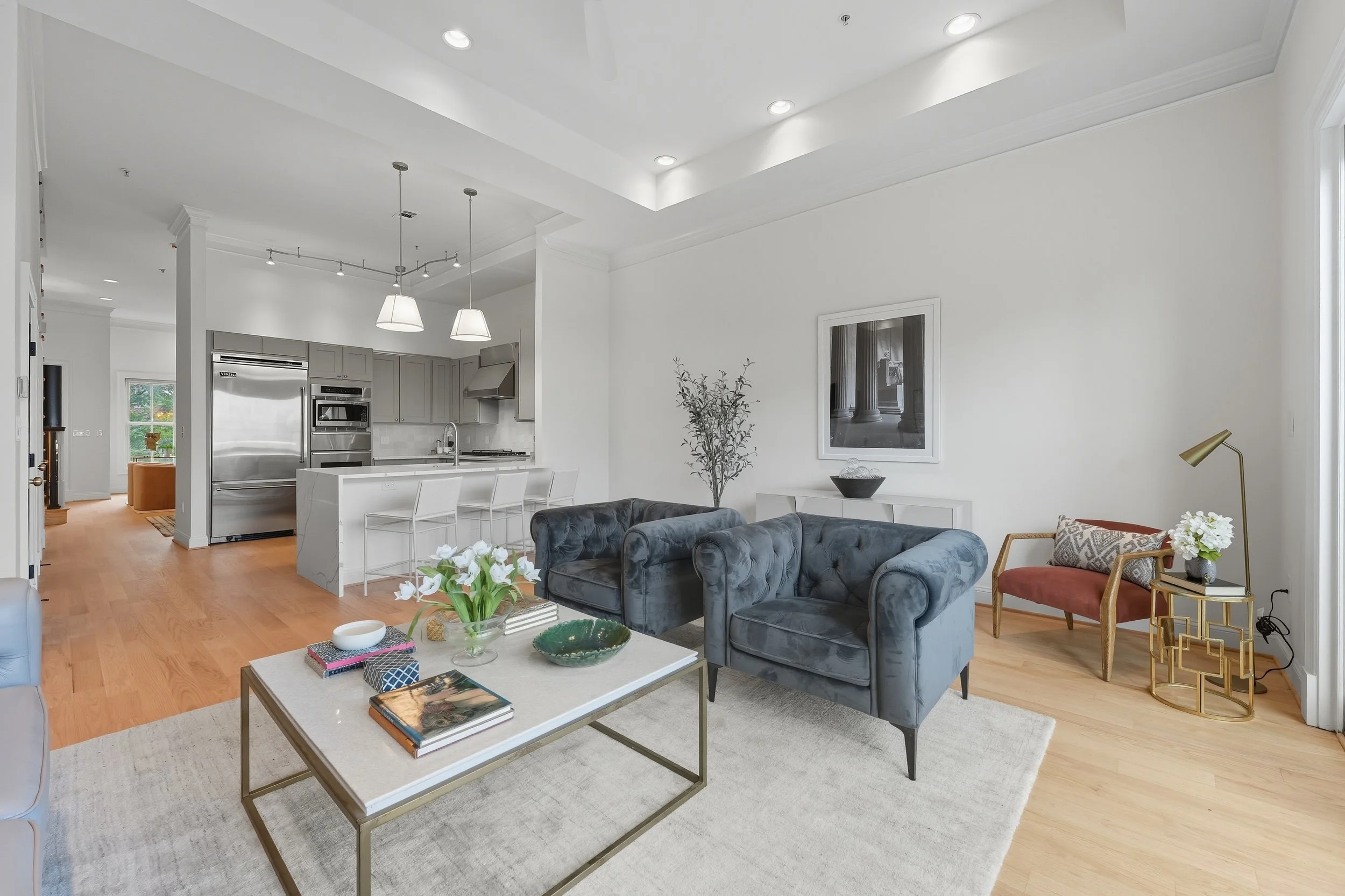 Modern living room with blue velvet tufted armchairs, a white coffee table with books and decorative items, and a side table with a gold frame and pink cushion. Open kitchen in background with stainless steel appliances and white cabinetry.