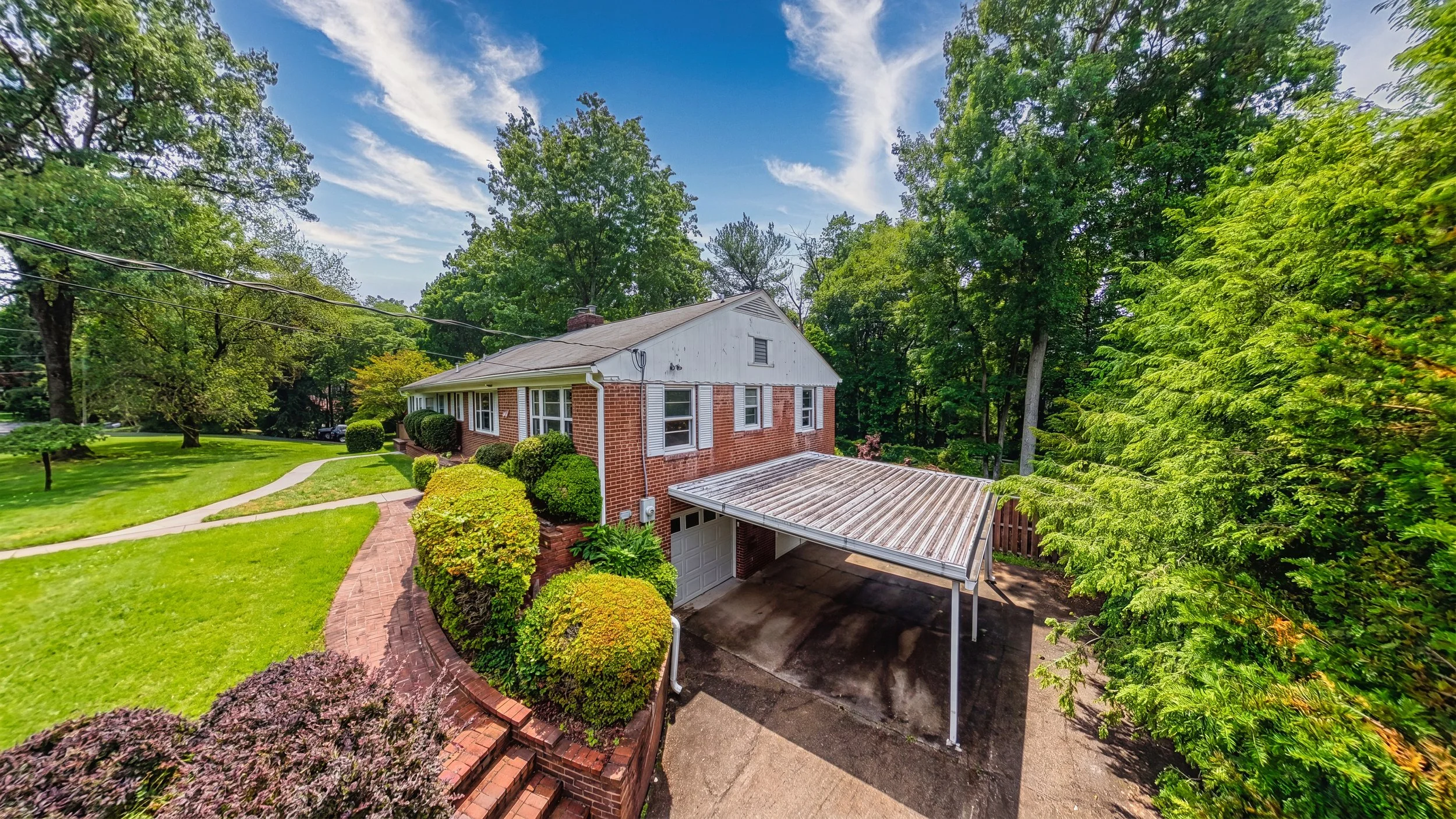 Elevated exterior view of a brick split-level with a carport, showing the driveway, front lawn, and mature landscaping from above eye level.