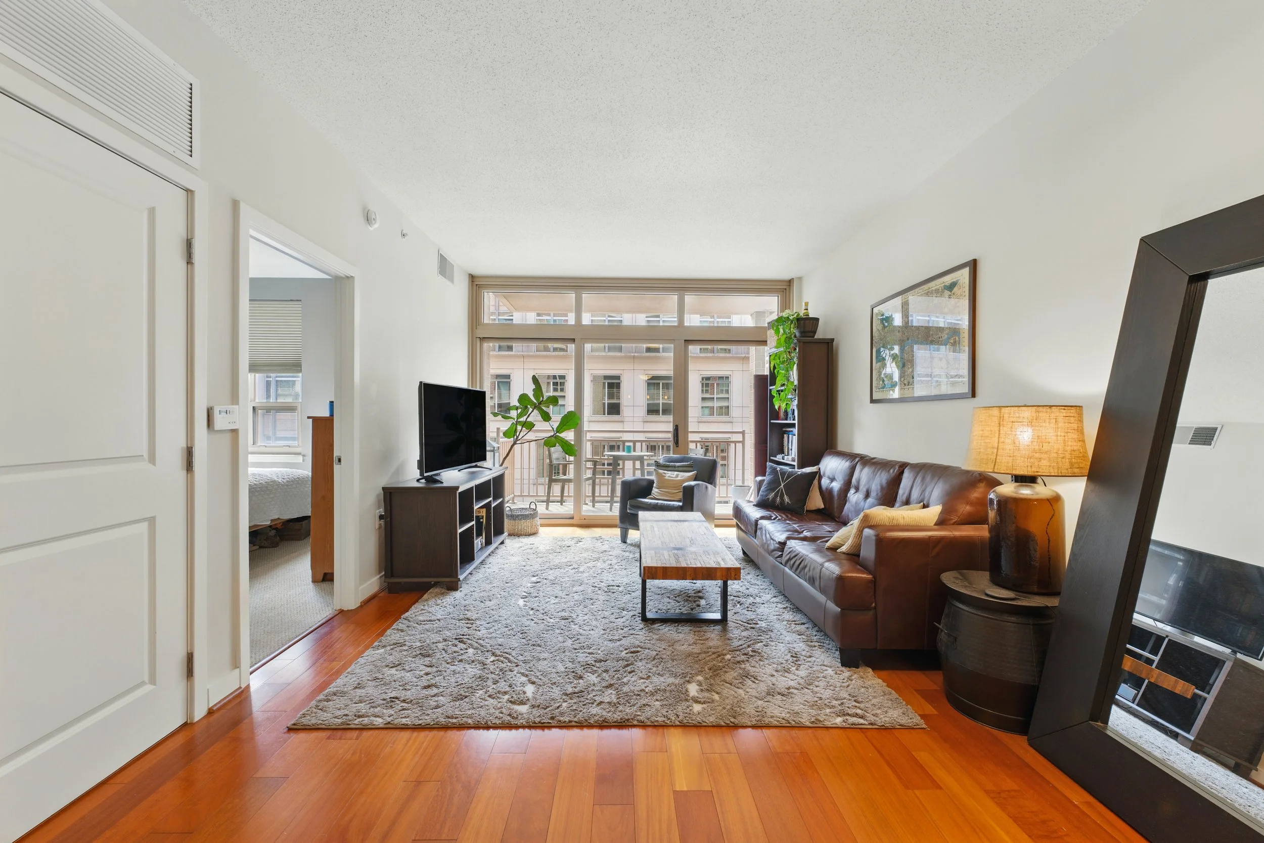 Living room with brown leather sofa, black and gray armchair, wooden coffee table, TV on a black stand, beige shag rug, large windows with balcony view, and a sliding door leading to a bedroom.