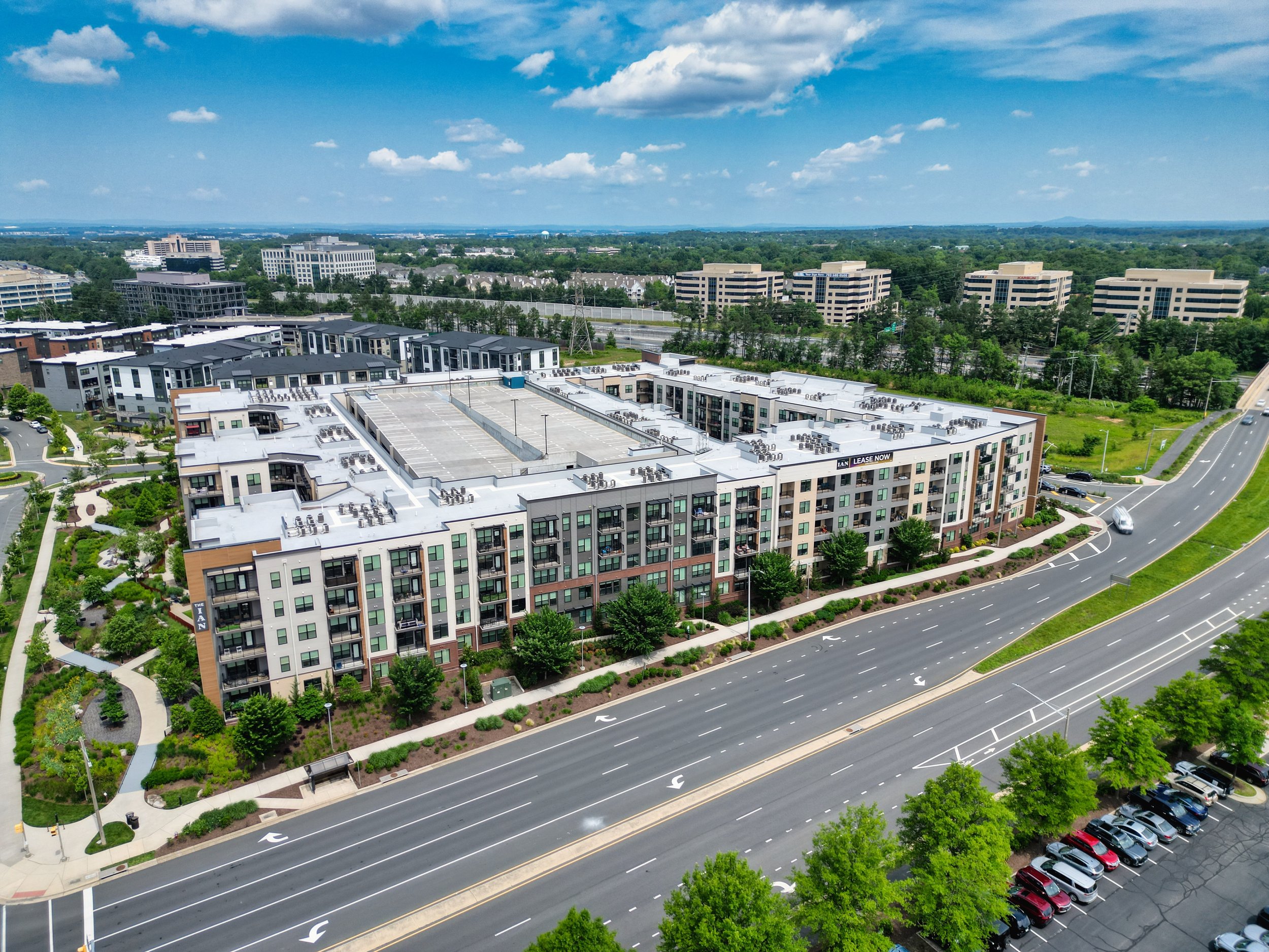 Aerial view of a modern multi-story apartment complex with landscaped gardens, parking lots, and surrounding streets, with office buildings and greenery in the background under a partly cloudy sky.