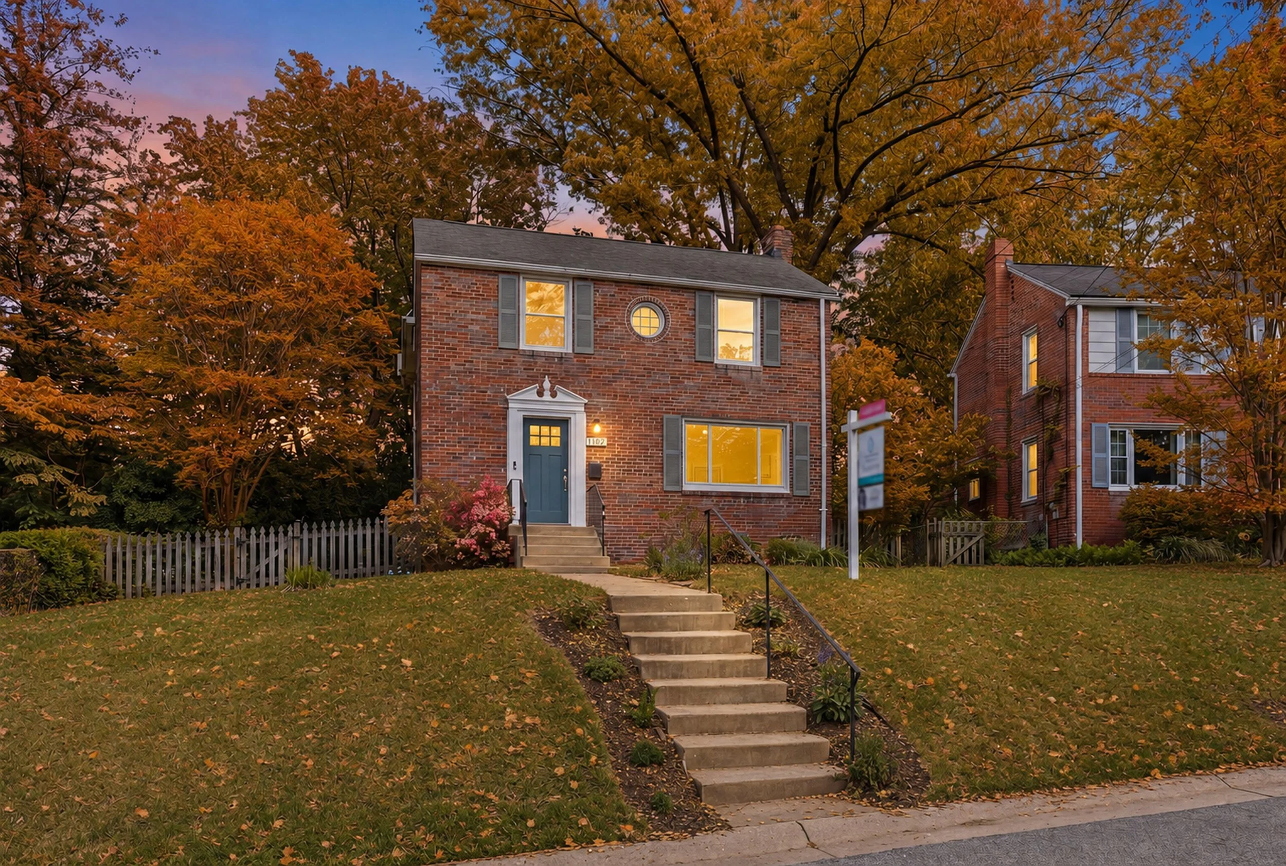 Two-story brick colonial at twilight with warm lit windows, a pink and blue sky, and orange fall canopy overhead.